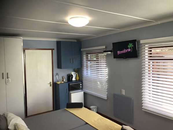 A compact bedroom with a bed, a small kitchenette, a TV, and a window with blinds. - 6 Stemmet Lodge (Hermanus)