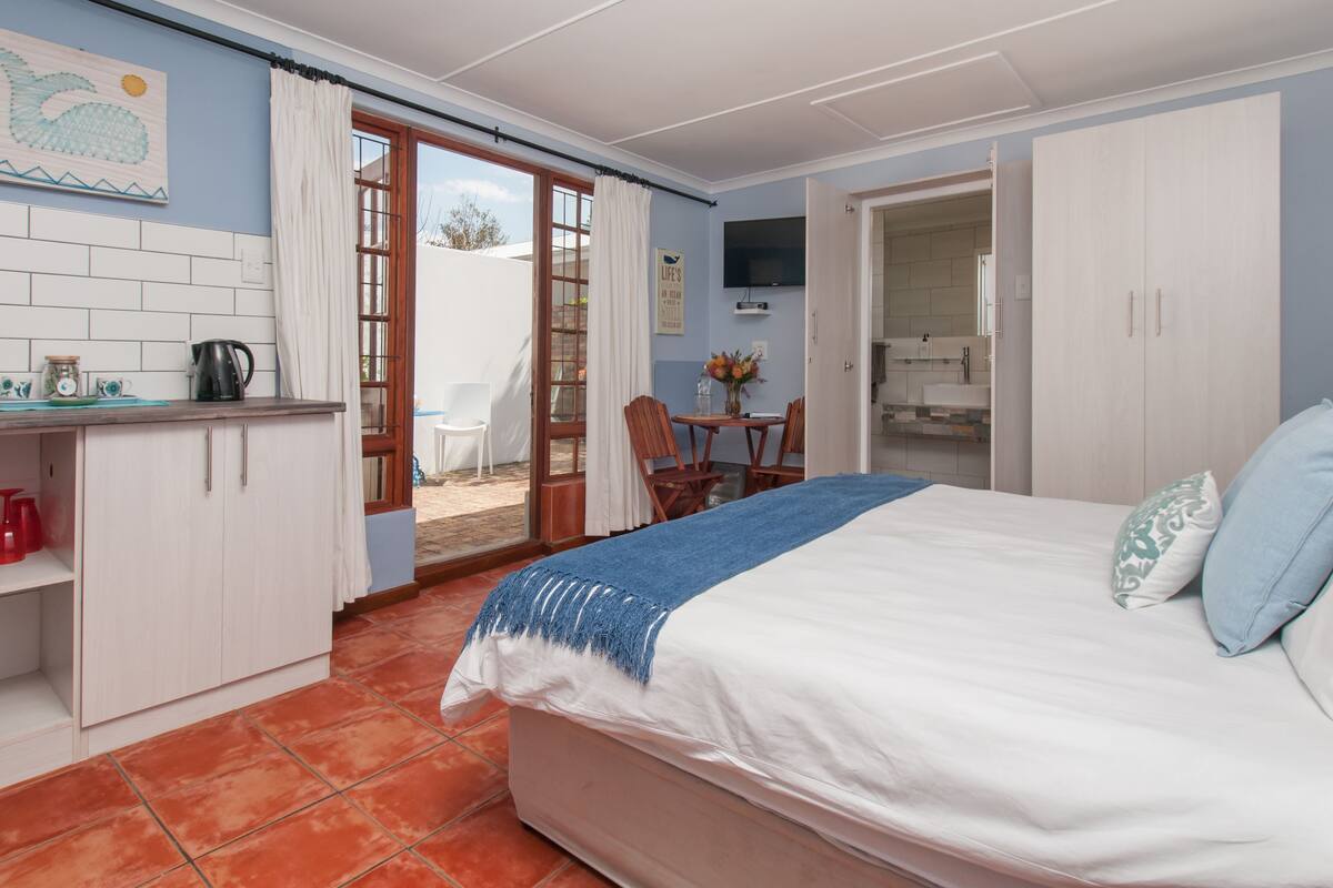 A bedroom with a bed, a desk with a kettle, a chair, and a small table with a vase.