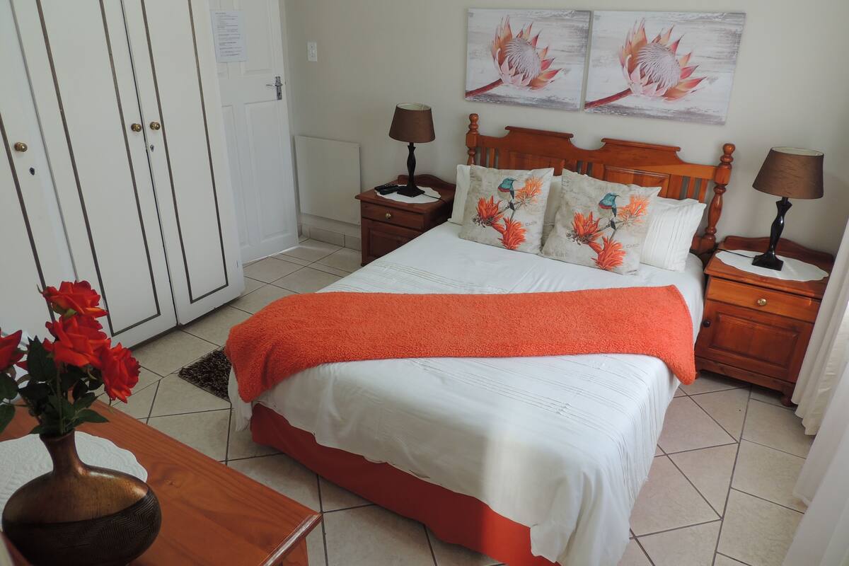 Ein ordentlich eingerichtetes Schlafzimmer mit einem Bett, zwei Nachttischen, einer hölzernen Kommode, einer Vase mit Blumen und zwei an der Wand befestigten Bildern.