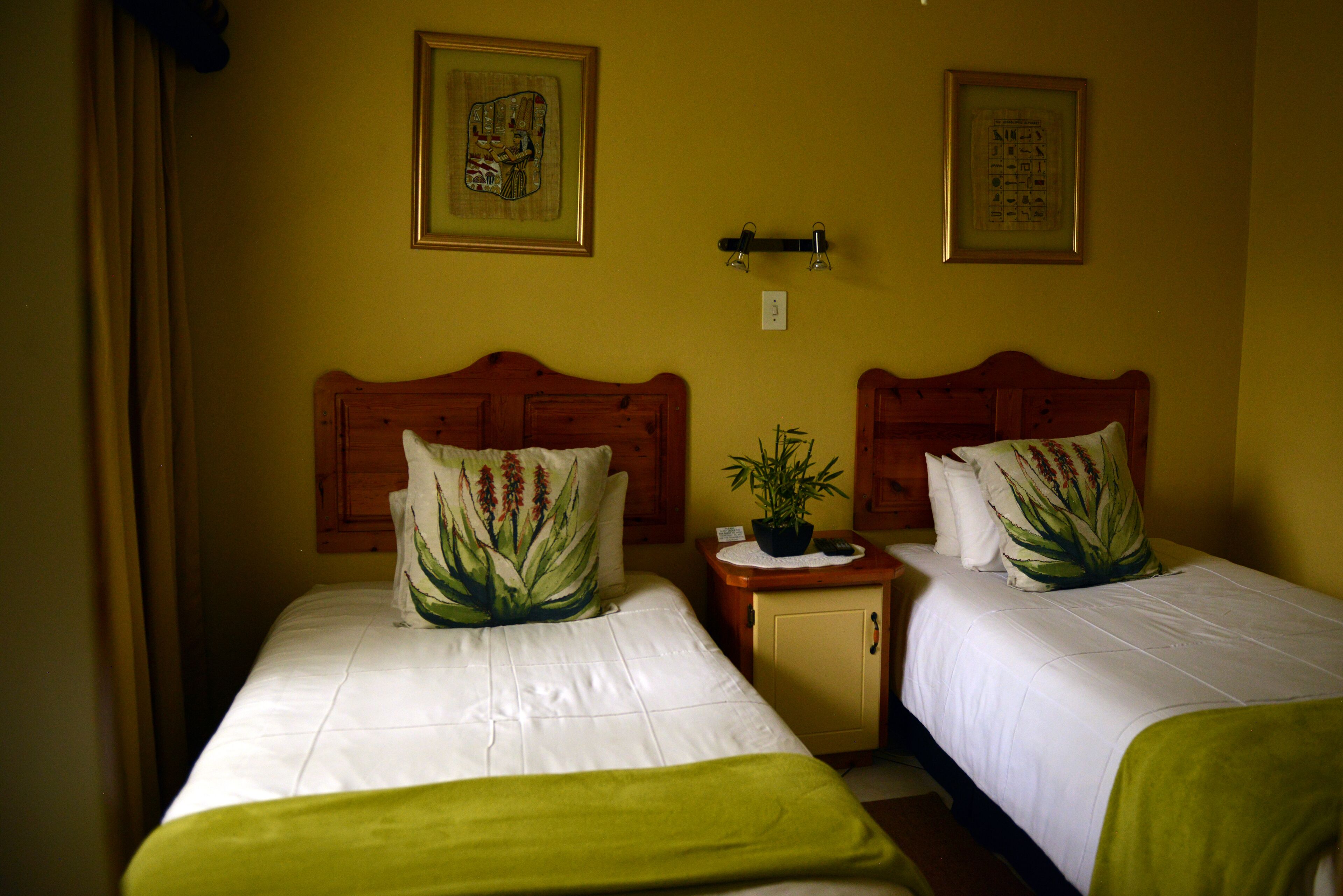 A hotel room with two single beds, each with a wooden headboard, white bedding, and green decorative pillows. There are two framed pictures on the wall above the beds.