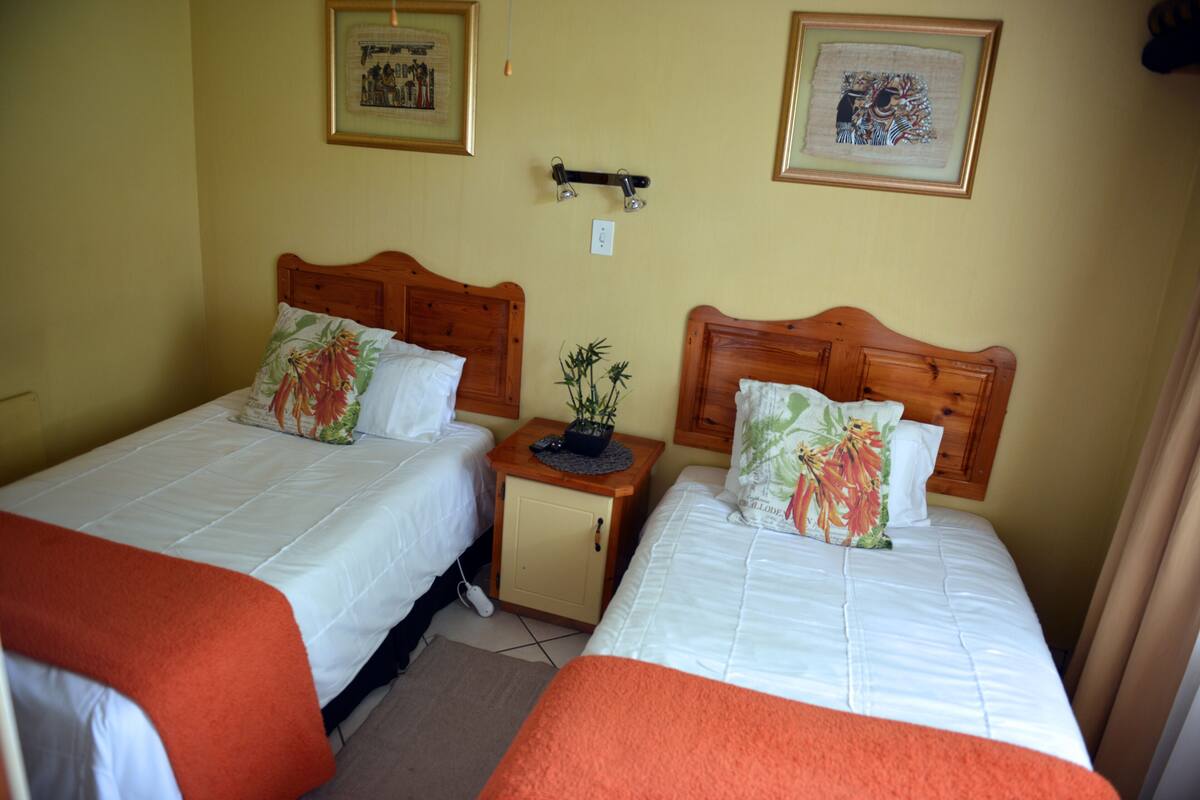 Ein Hotelzimmer mit zwei Einzelbetten, jeweils mit einem hölzernen Kopfteil, weißer Bettwäsche und einer roten Tagesdecke. Zwischen den Betten befindet sich ein kleiner Nachttisch mit einer Pflanze, und über jedem Bett hängt eine Wandleuchte.
