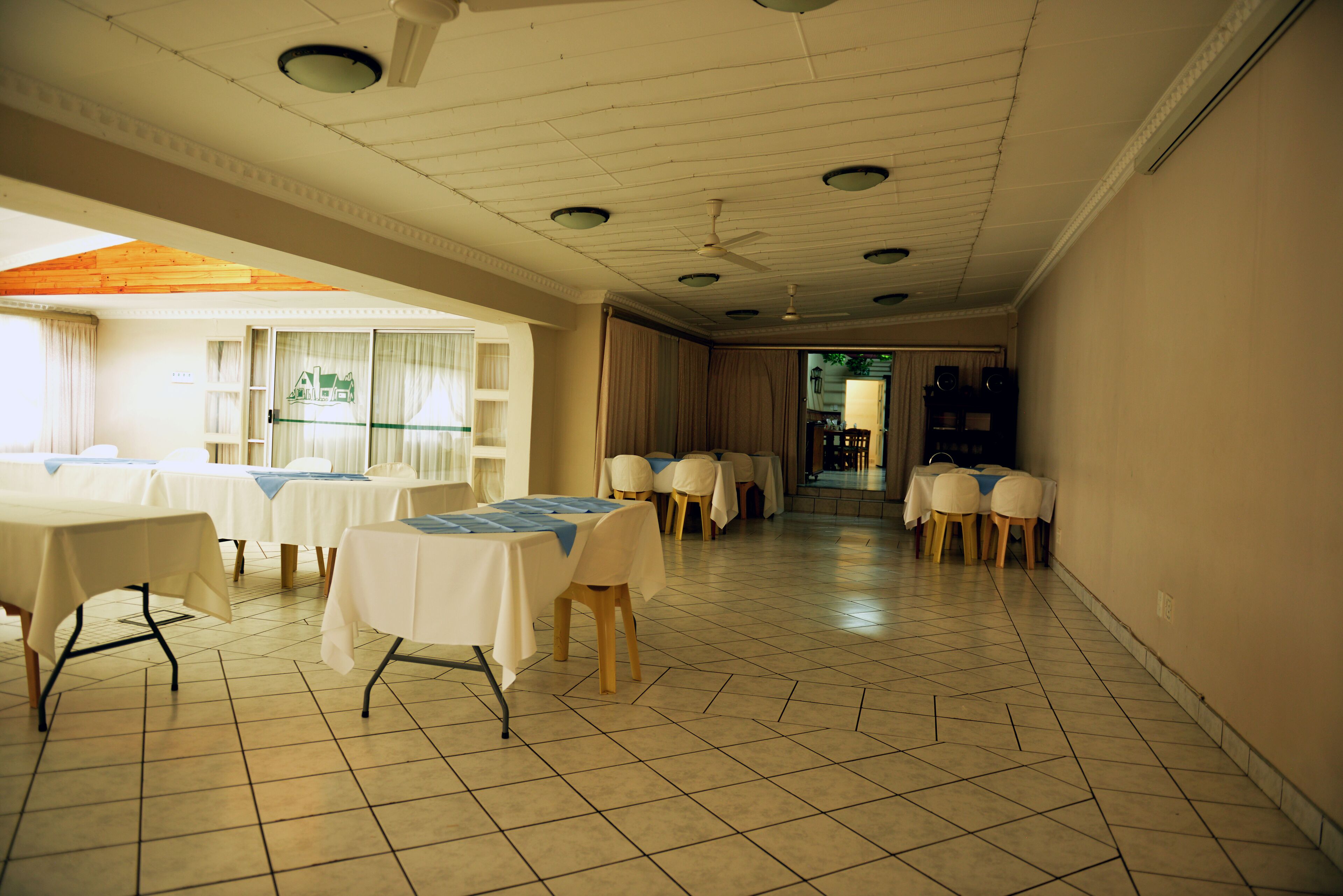 A hotel dining area with multiple tables and chairs, a tiled floor, and ceiling fans.
