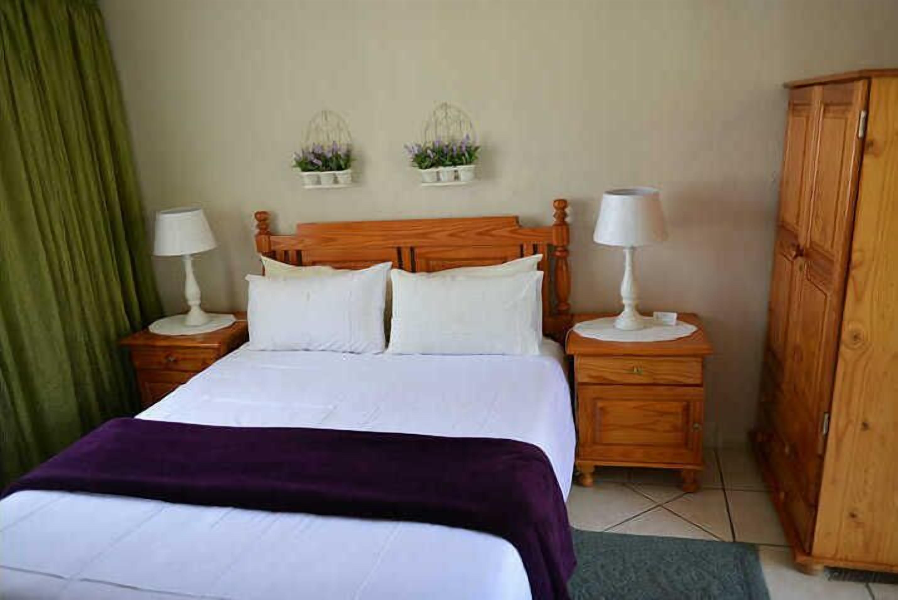A neatly made bed with white linens and a purple throw blanket, flanked by wooden nightstands with lamps, a green curtain, and a wooden wardrobe.