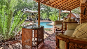 An outdoor seating area with wicker furniture, a glass-top table, and a pool in the background. - Kruger Riverside Lodge (Nkomazi)