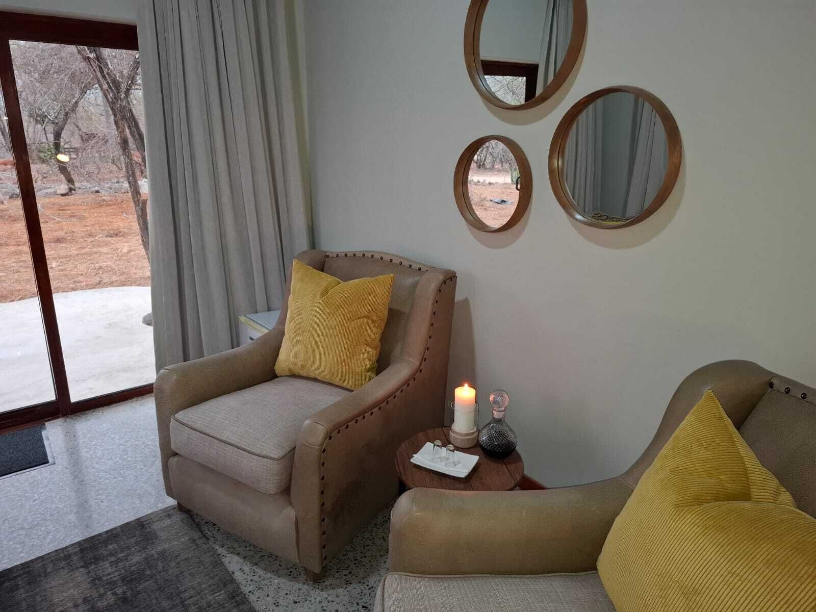 Una habitación con un sillón y un sofá color beige, ambos con cojines amarillos. Hay tres espejos redondos en la pared, una vela encendida sobre una mesita y una vista de un paisaje nevado a través de una puerta de cristal.