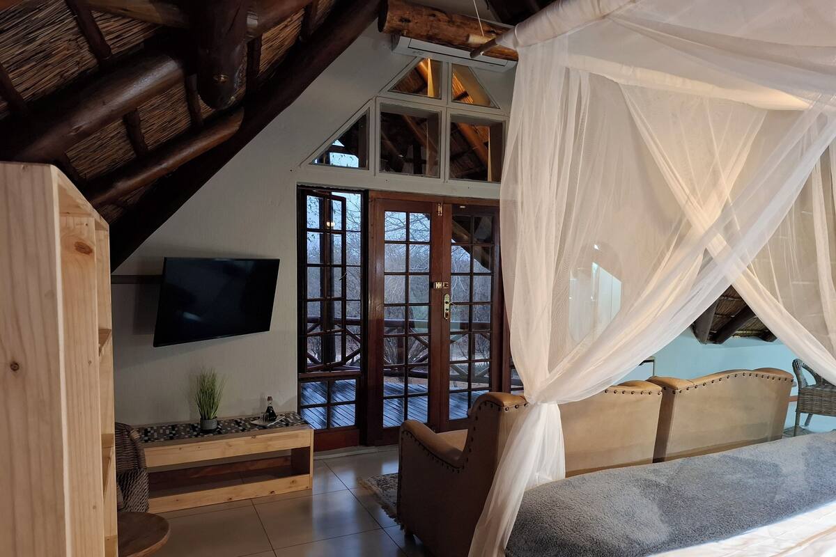 Quarto com cama, televisão, um banco de madeira e uma grande janela com vista para o exterior.