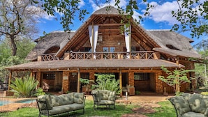 A rustic lodge with a thatched roof, a covered patio with outdoor seating, and a swimming pool. - Kruger Riverside Lodge (Nkomazi)