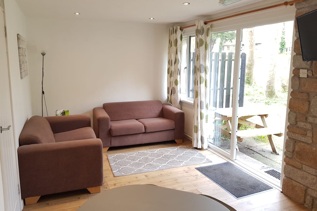 Ein gemütliches Wohnzimmer mit einer braunen Couch, einem kleineren Sessel und einem Couchtisch. Auf dem Boden liegt ein gemustertes Teppich und eine Tür führt in einen Außenbereich mit einem Holztisch und Stühlen.