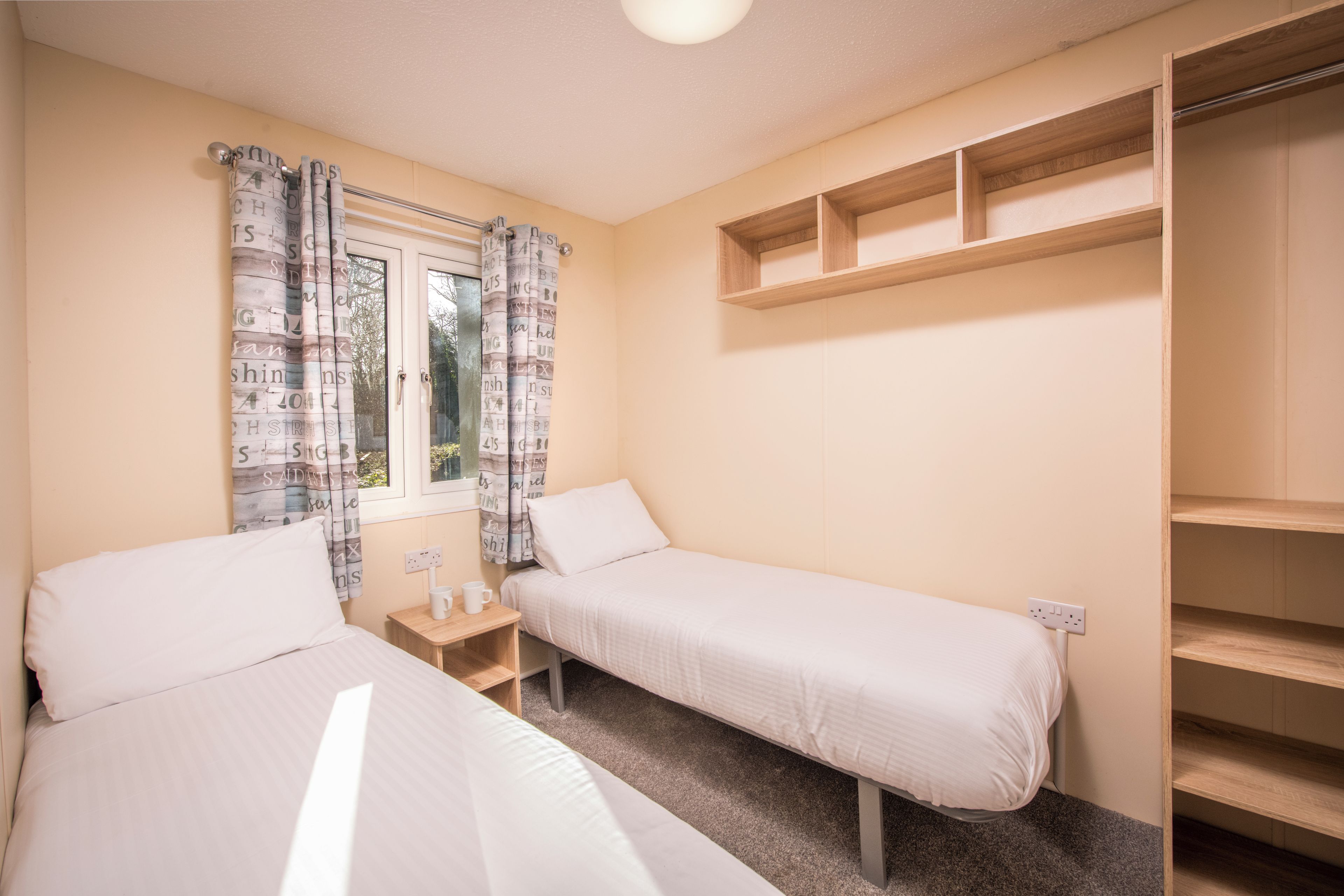 Een slaapkamer met twee eenpersoonsbedden, een klein nachtkastje met een lamp, een raam met gordijnen en een houten plank.