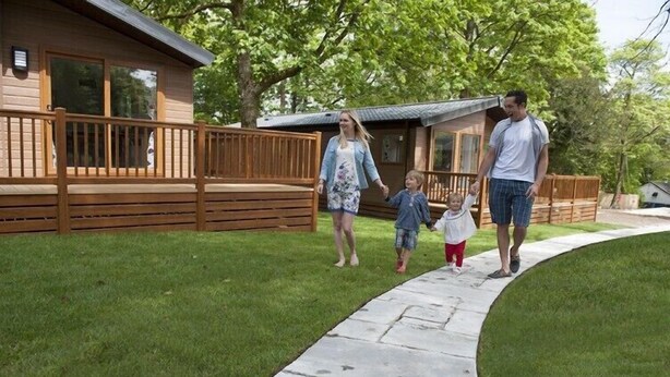A family walking on a stone pathway towards a wooden decked house with a grassy yard. - St Ives Holiday Village (St Ives)