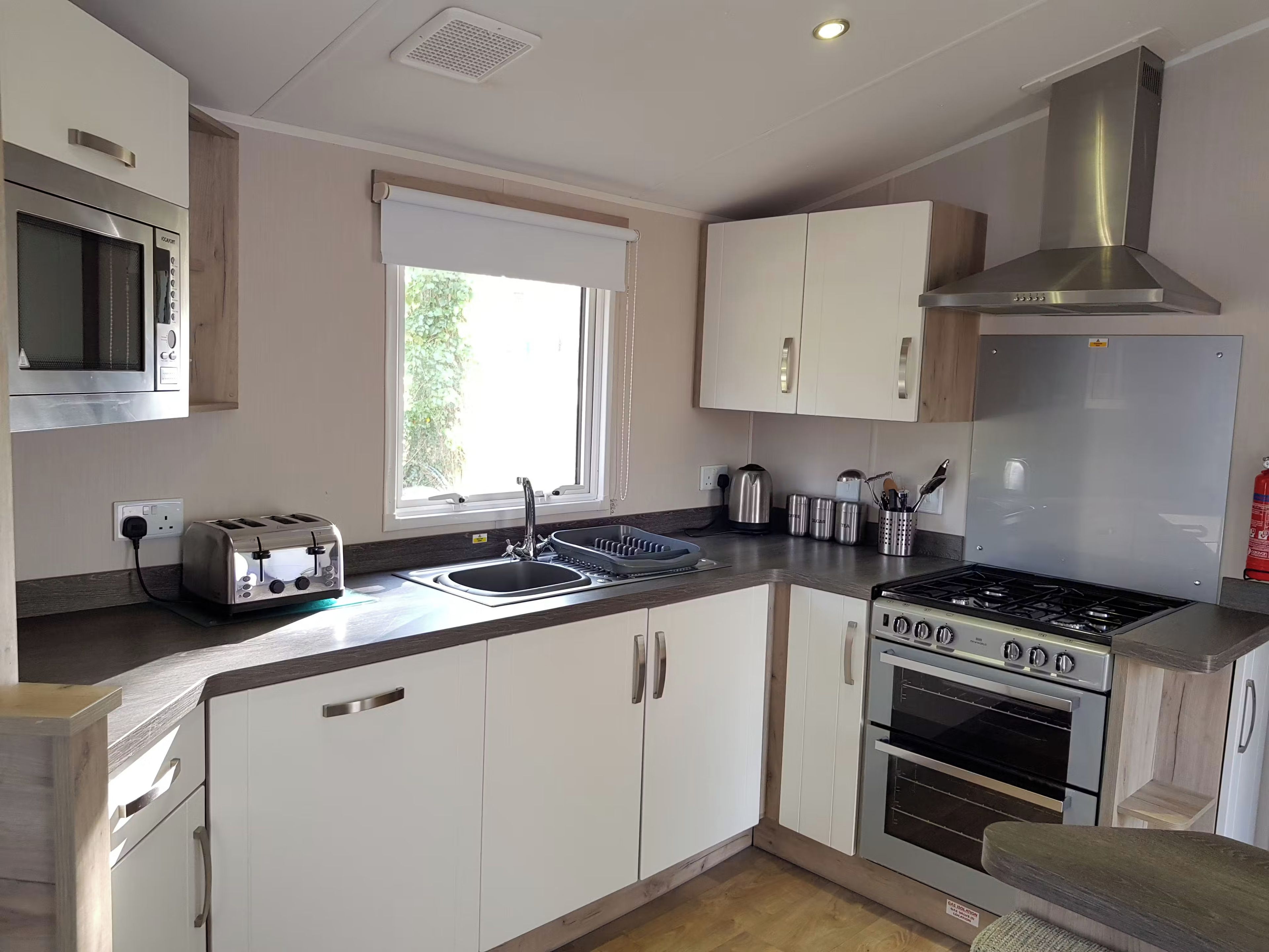A modern kitchen with white cabinets, a stainless steel range hood, a built-in oven, a microwave, and a window providing natural light.