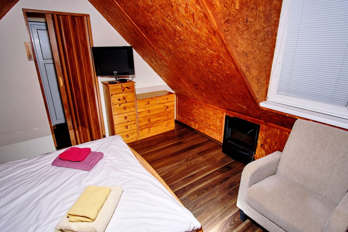 Een slaapkamer met een schuine plafond, houten meubels, een bed met handdoeken, een televisie op een dressoir en een raam met jaloezieën.