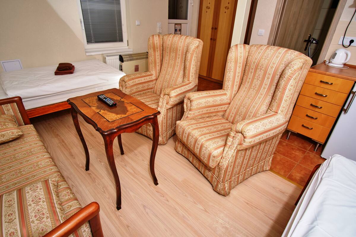 Een hotelkamer met een bed, twee gestreepte fauteuils, een houten tafel met een afstandsbediening, een kleine tafellamp, een houten kast en een radiator.