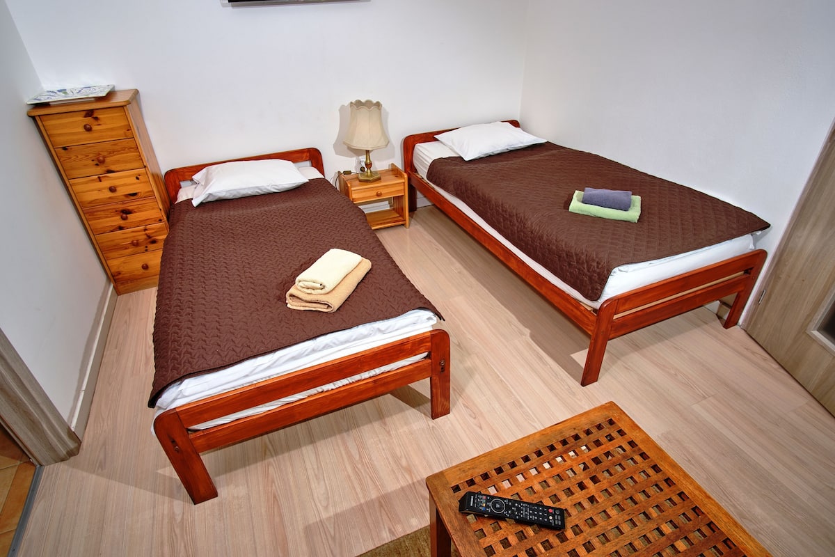 Twee houten bedden met bruin beddengoed, een houten dressoir, een nachtkastje met een lamp en een houten tafel met een afstandsbediening.