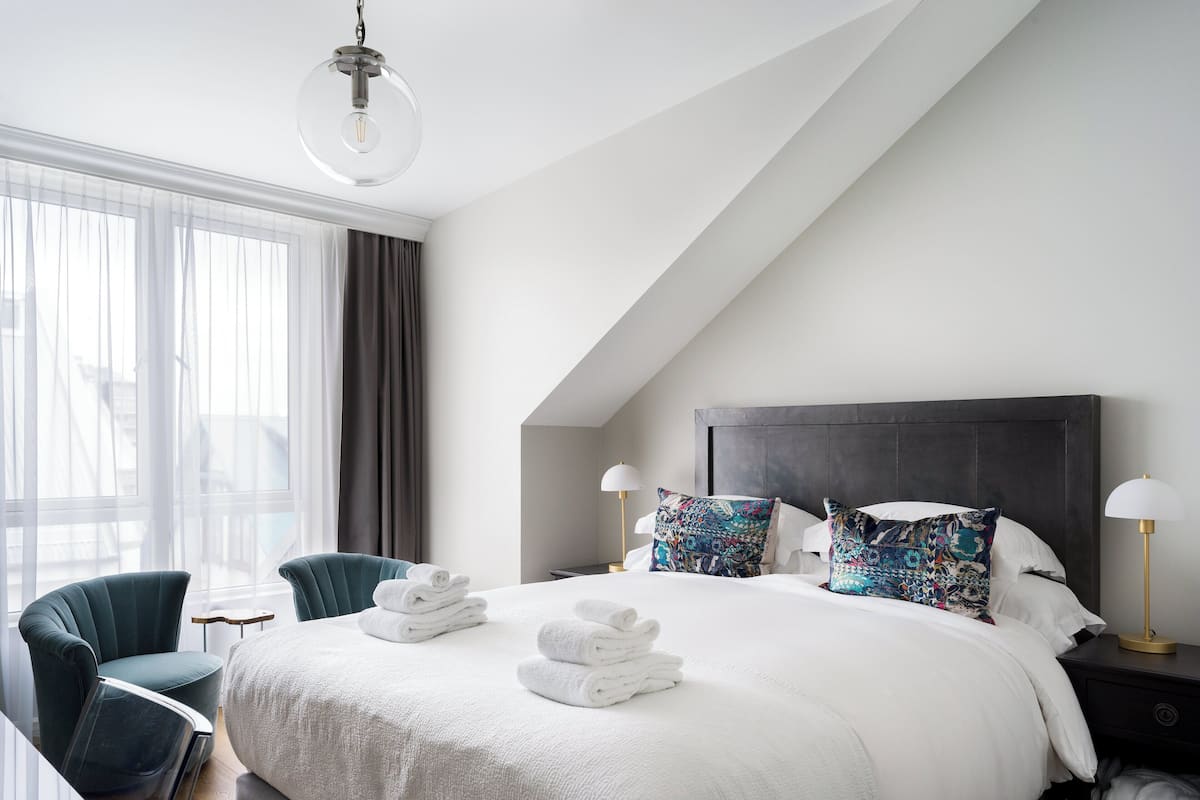 Una habitación de hotel moderna con una cama grande, dos mesitas de noche, un escritorio, una silla y un amplio ventanal con cortinas.