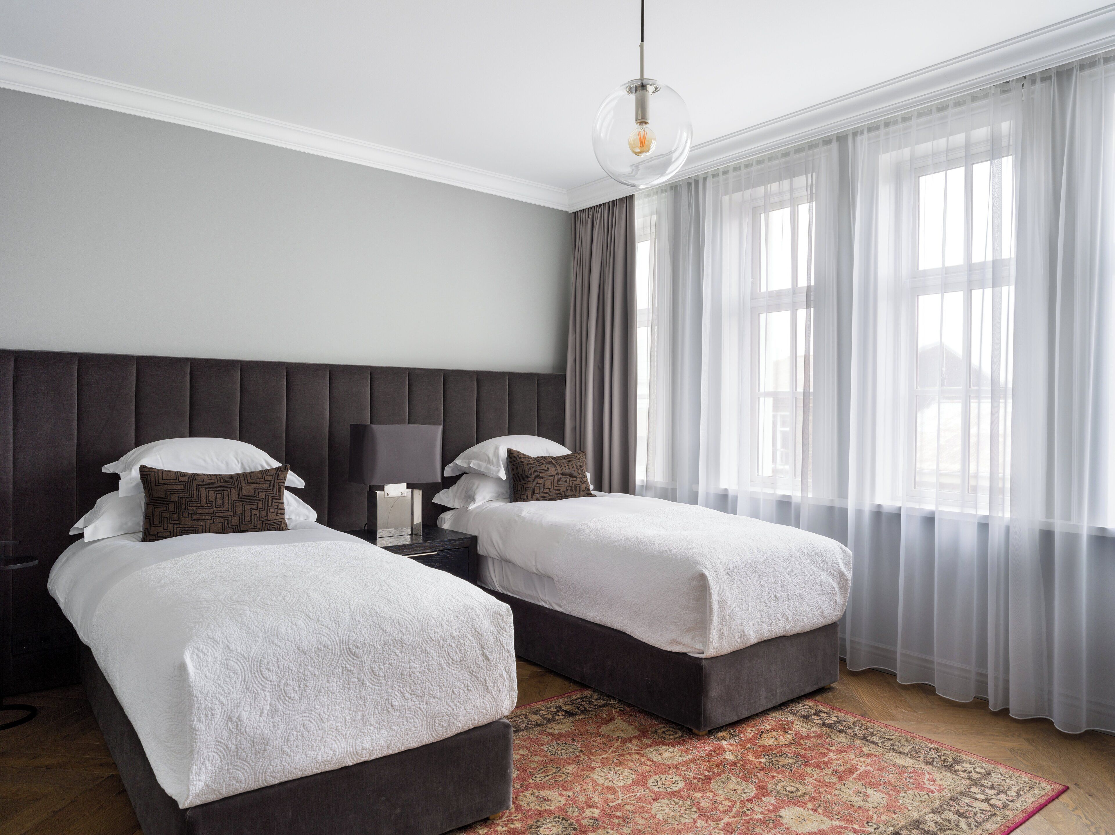 A hotel room with two beds, a nightstand with a lamp, a large window with sheer curtains, and a patterned rug on the floor.