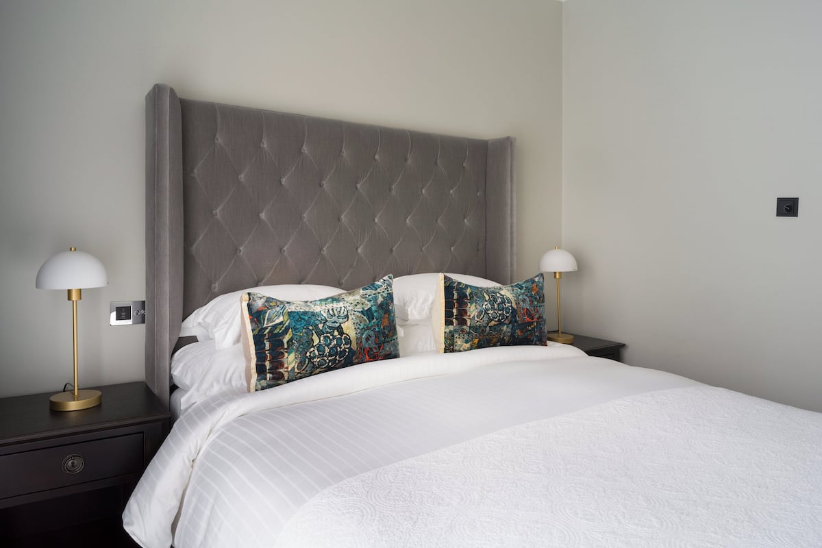 Un lit bien fait, avec une tête de lit rembourrée de couleur grise, du linge de lit blanc et deux coussins décoratifs.