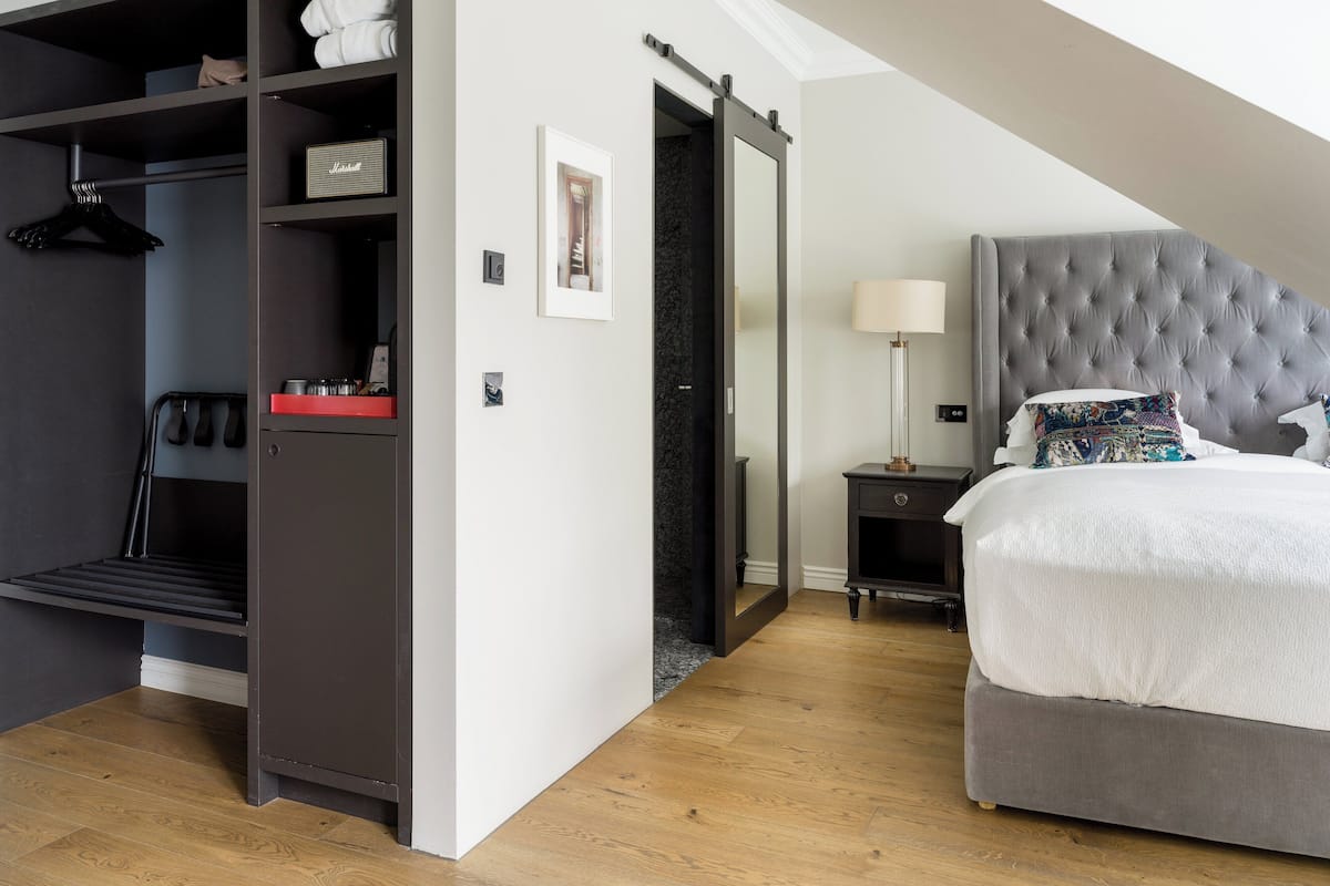 Une chambre d’hôtel moderne avec une tête de lit rembourrée de couleur grise, un grand lit avec une literie blanche, une table de chevet avec une lampe, une armoire contenant des vêtements suspendus et un tableau encadré au mur.