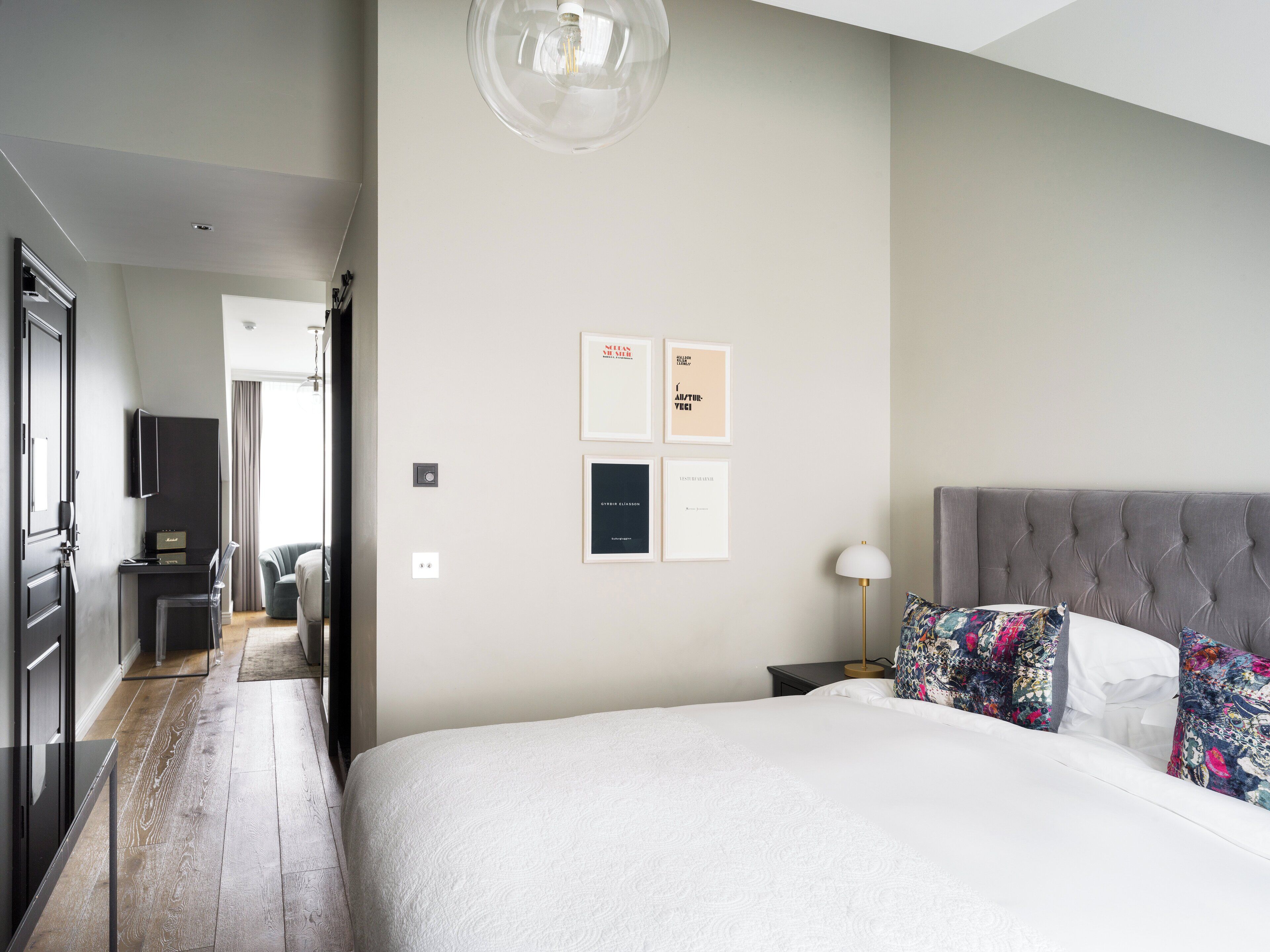 Un dormitorio moderno con una cama grande, un cabecero tapizado en gris y una mesita de noche con una lámpara. Hay cuadros enmarcados en la pared y un pasillo que lleva a otra habitación.