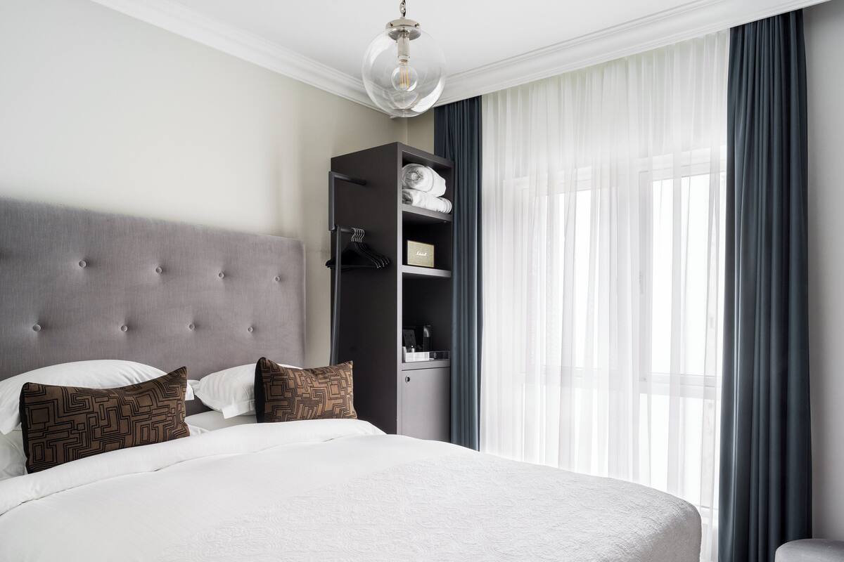 Una cama bien tendida con cabecera gris, un armario colgado y un ventanal con cortinas transparentes.