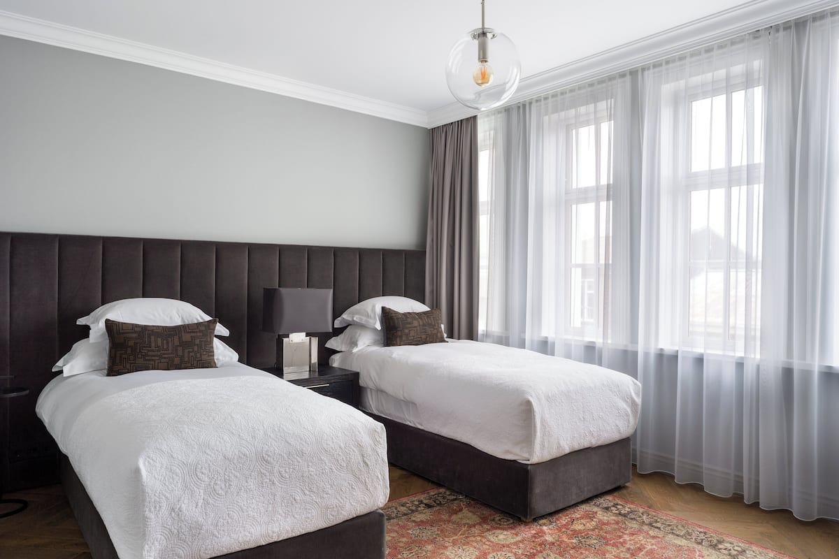 Una habitación de hotel con dos camas, una mesita de noche con lámpara, un ventanal con cortinas traslúcidas y una alfombra estampada en el suelo.