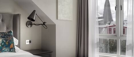 A bedroom with a bed, a nightstand, a lamp, a window with a view of a church, and a painting on the wall.