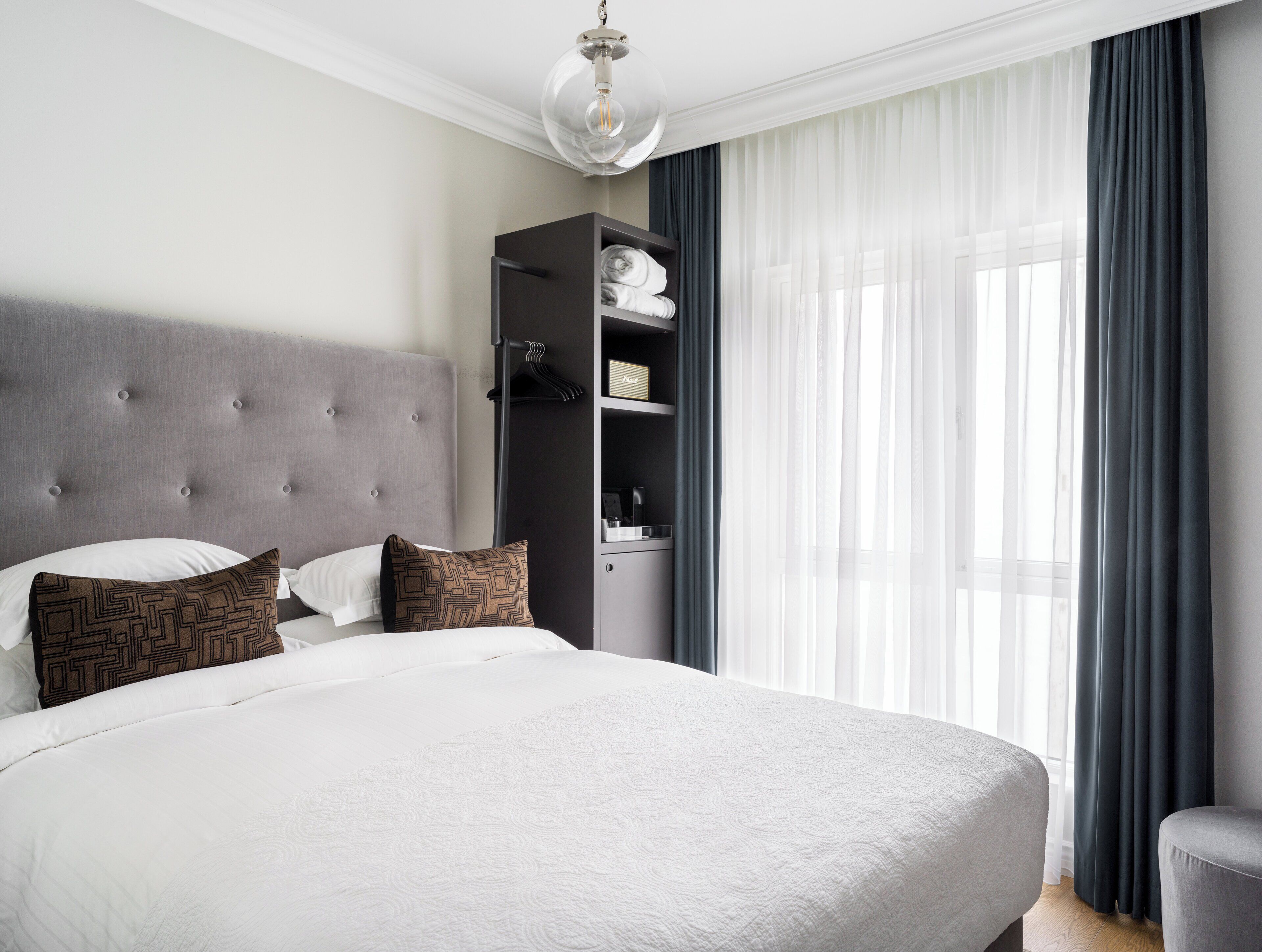 Uma cama bem arrumada com cabeceira cinza, um guarda-roupa suspenso e uma janela grande com cortinas leves.