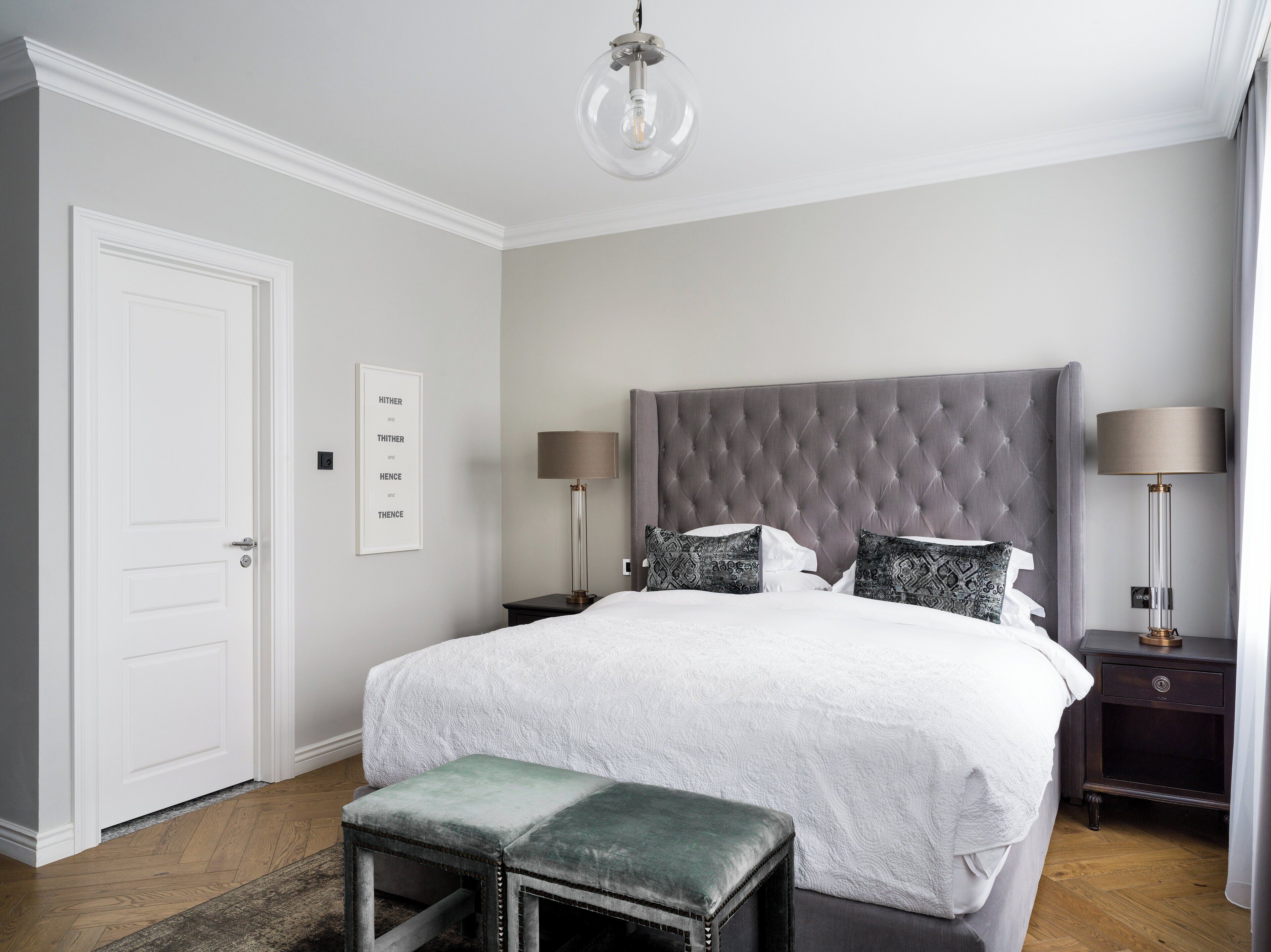 A bedroom with a tufted headboard, a large bed with white linens, two bedside tables with lamps, a bench, and a door.