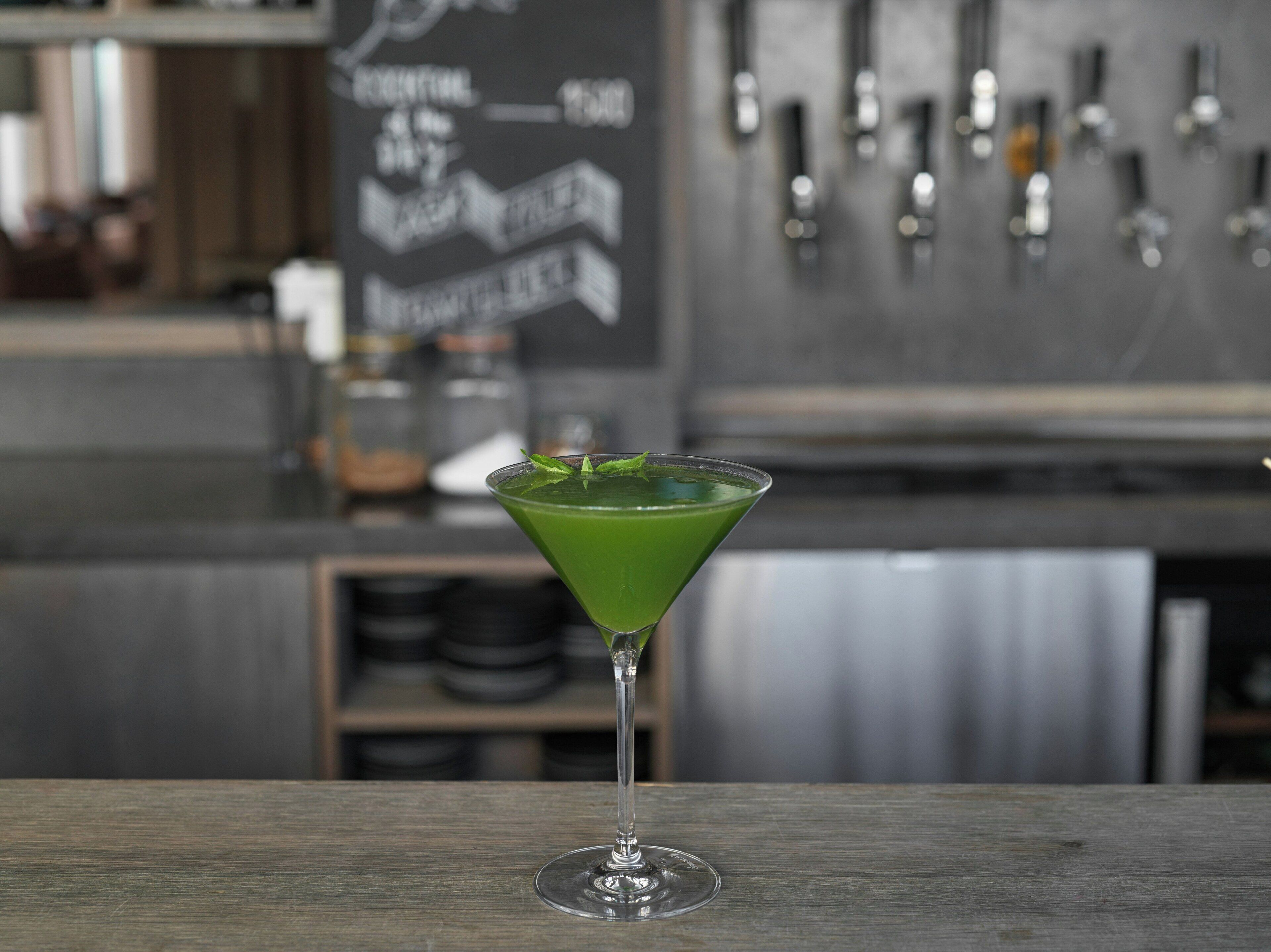 A green cocktail in a martini glass on a wooden surface with a bar and shelves in the background.