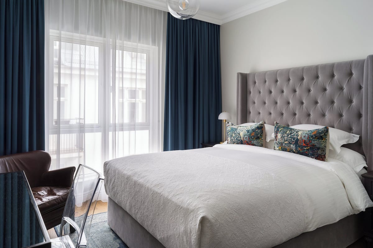 Habitación de hotel moderna con una cama grande, cabecera capitonada y vistas de la ciudad a través de cortinas traslúcidas.