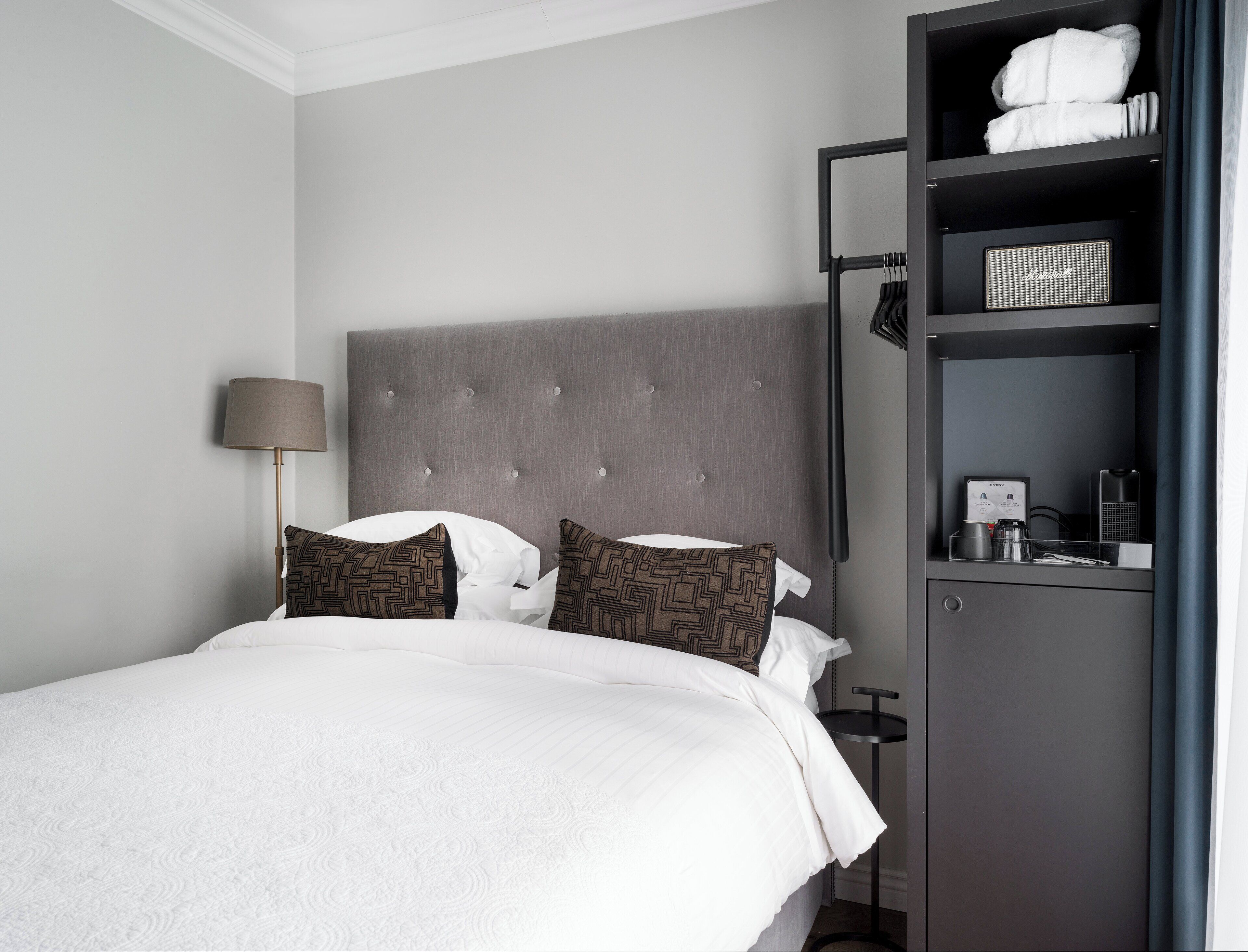 A neatly made bed with white linens and patterned pillows, a grey upholstered headboard, a bedside table with a lamp, and a built-in shelf with towels and toiletries.