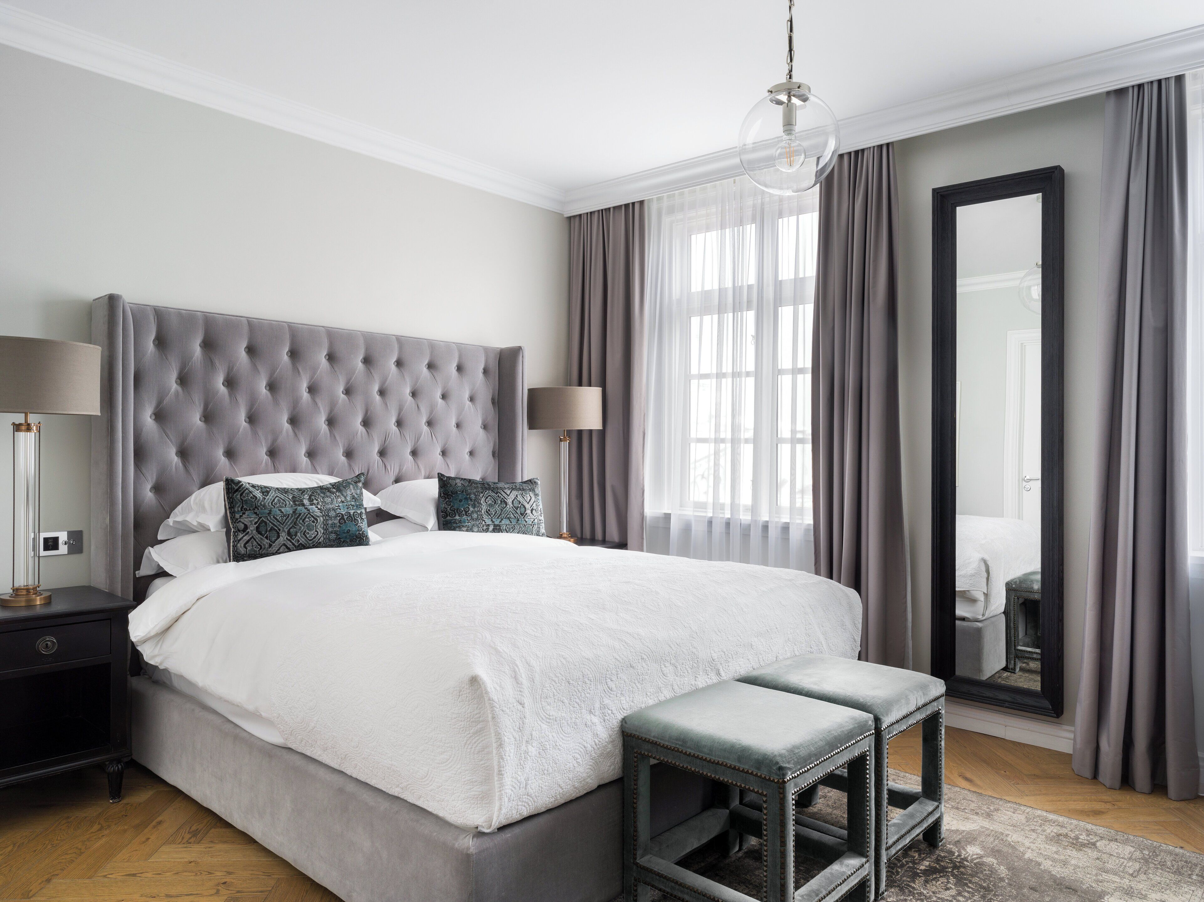 A neatly made bed with a tufted headboard, two bedside tables with lamps, a bench, and a large mirror.