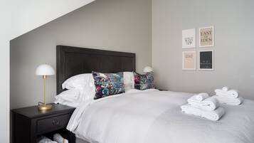 Una cama bien hecha con sábanas blancas, cabecera de madera oscura y una mesita de noche con una lámpara. Hay toallas apiladas sobre la cama y un pequeño estante con toallas adicionales.