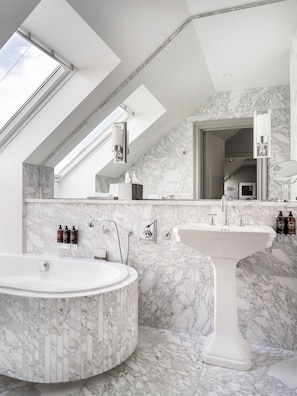 A modern bathroom with a skylight, a freestanding bathtub, a pedestal sink, and marble walls.