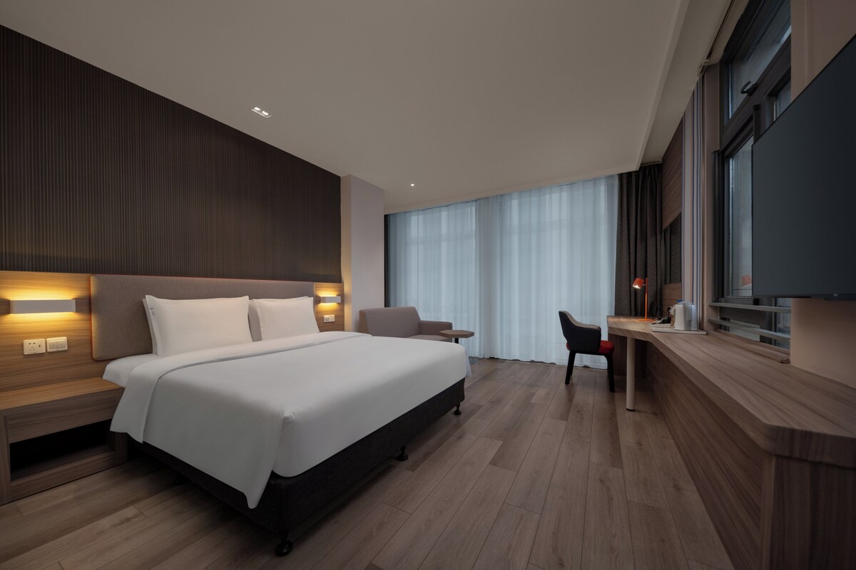 A modern hotel room with a large bed, bedside tables, a desk, a chair, and a TV.