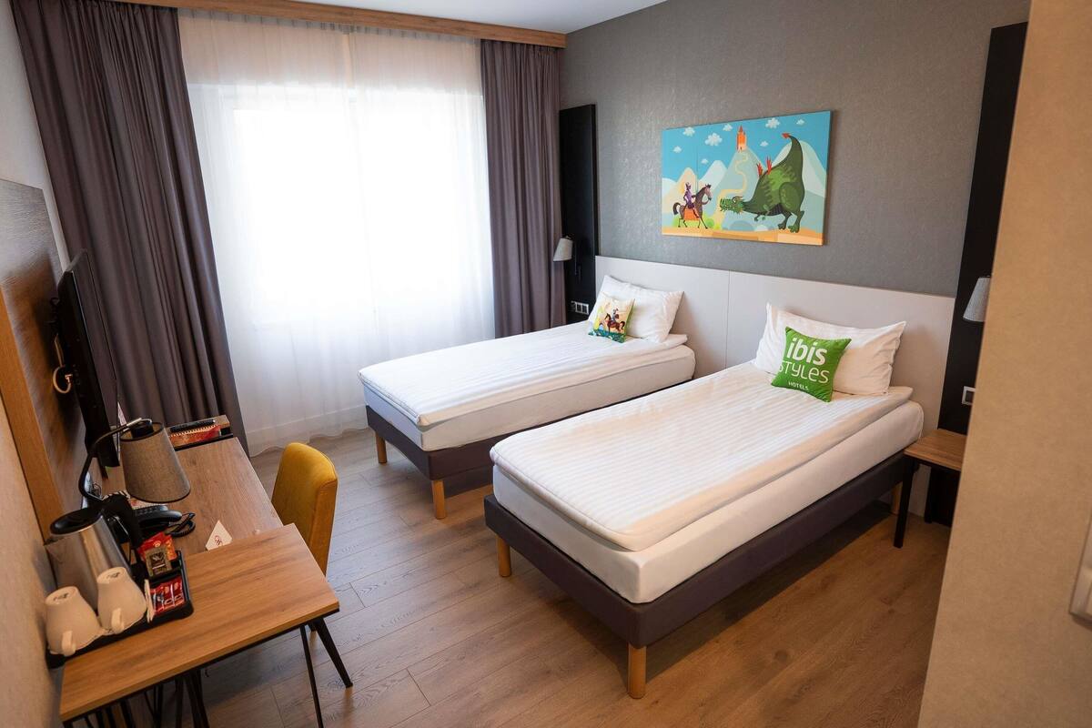 A hotel room with two beds, a desk with a coffee maker, a TV, and a painting on the wall.