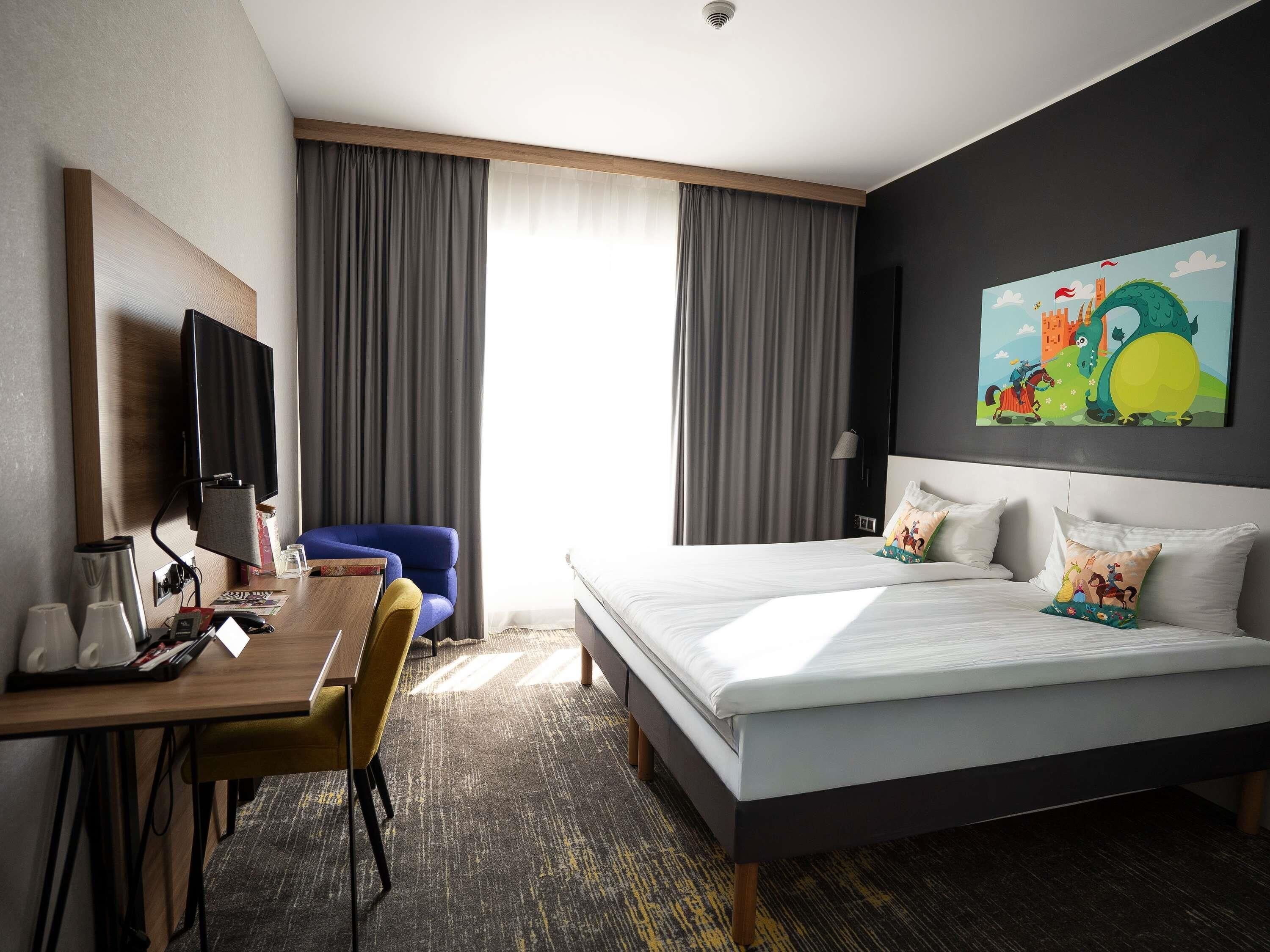 A hotel room with a bed, a desk, a chair, a TV, and a painting on the wall.