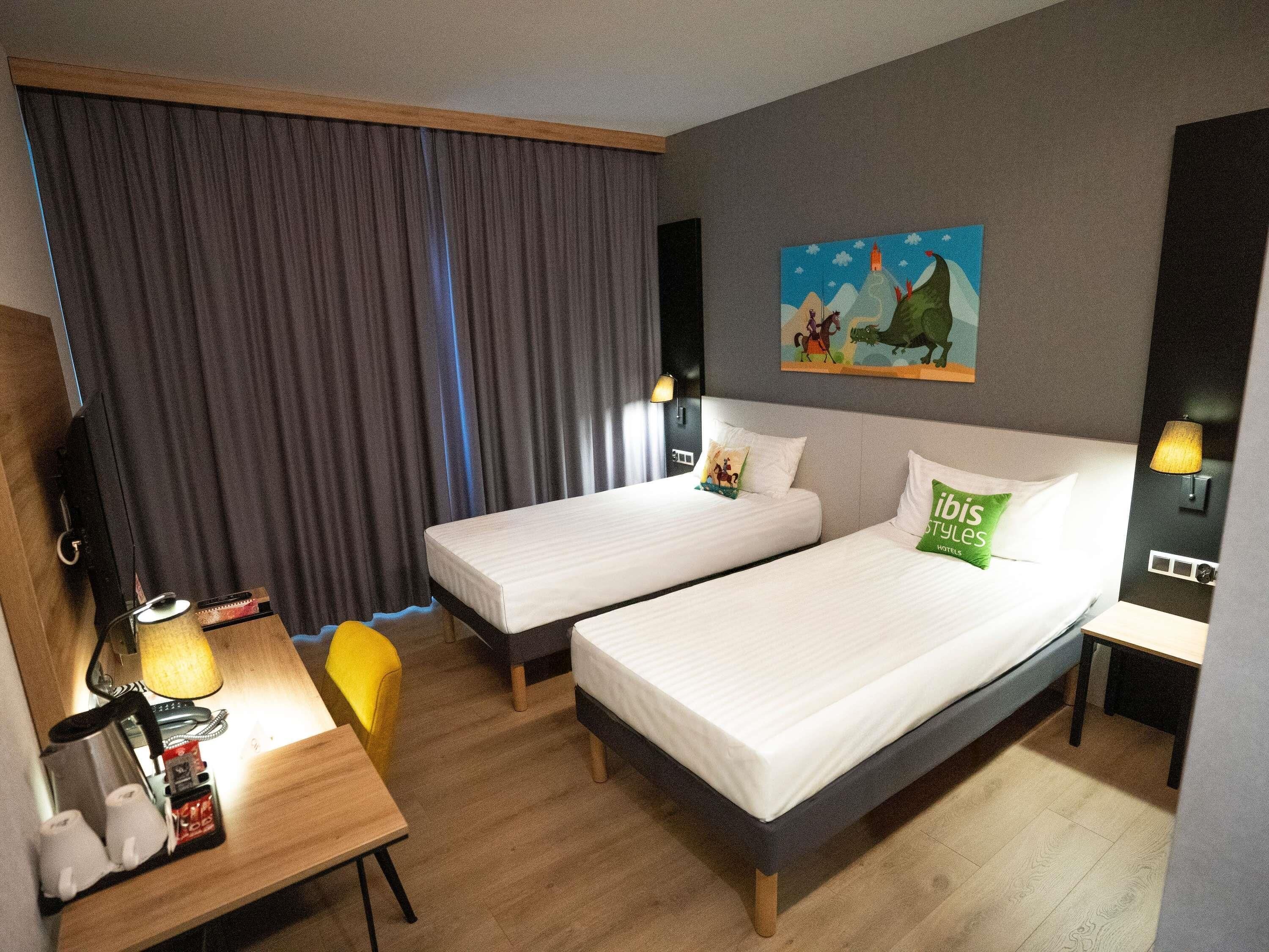 A hotel room with two beds, a desk with a lamp, a chair, and a painting on the wall.