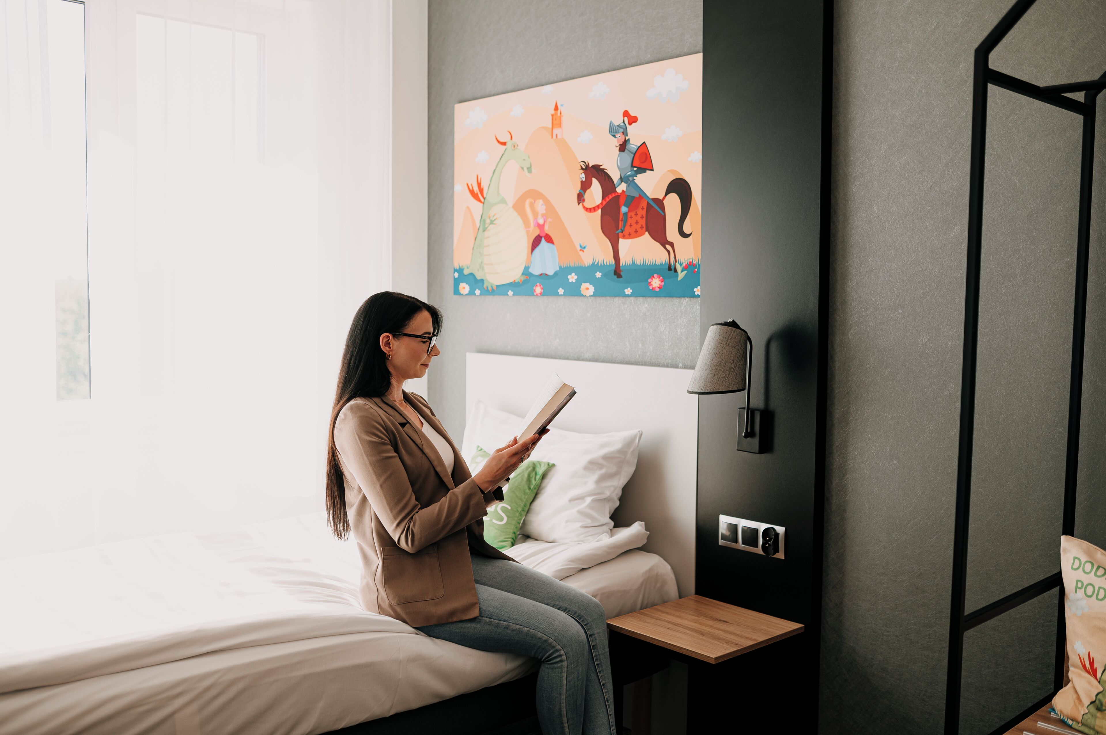 A person sitting on a bed in a room with a painting on the wall, a bedside table, and a lamp.