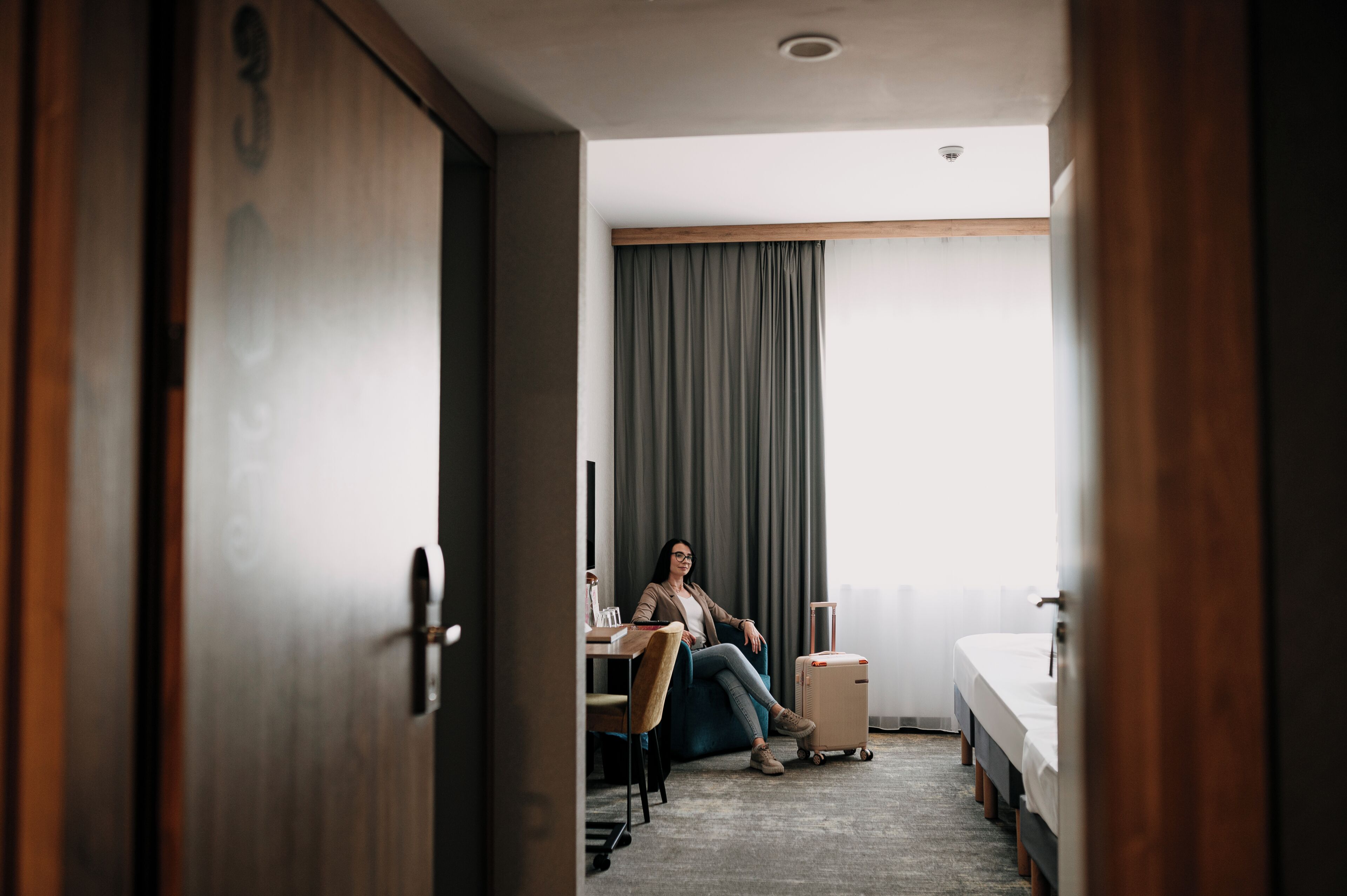 Une chambre d’hôtel avec un lit, un bureau, une chaise, une valise et une fenêtre avec des rideaux.