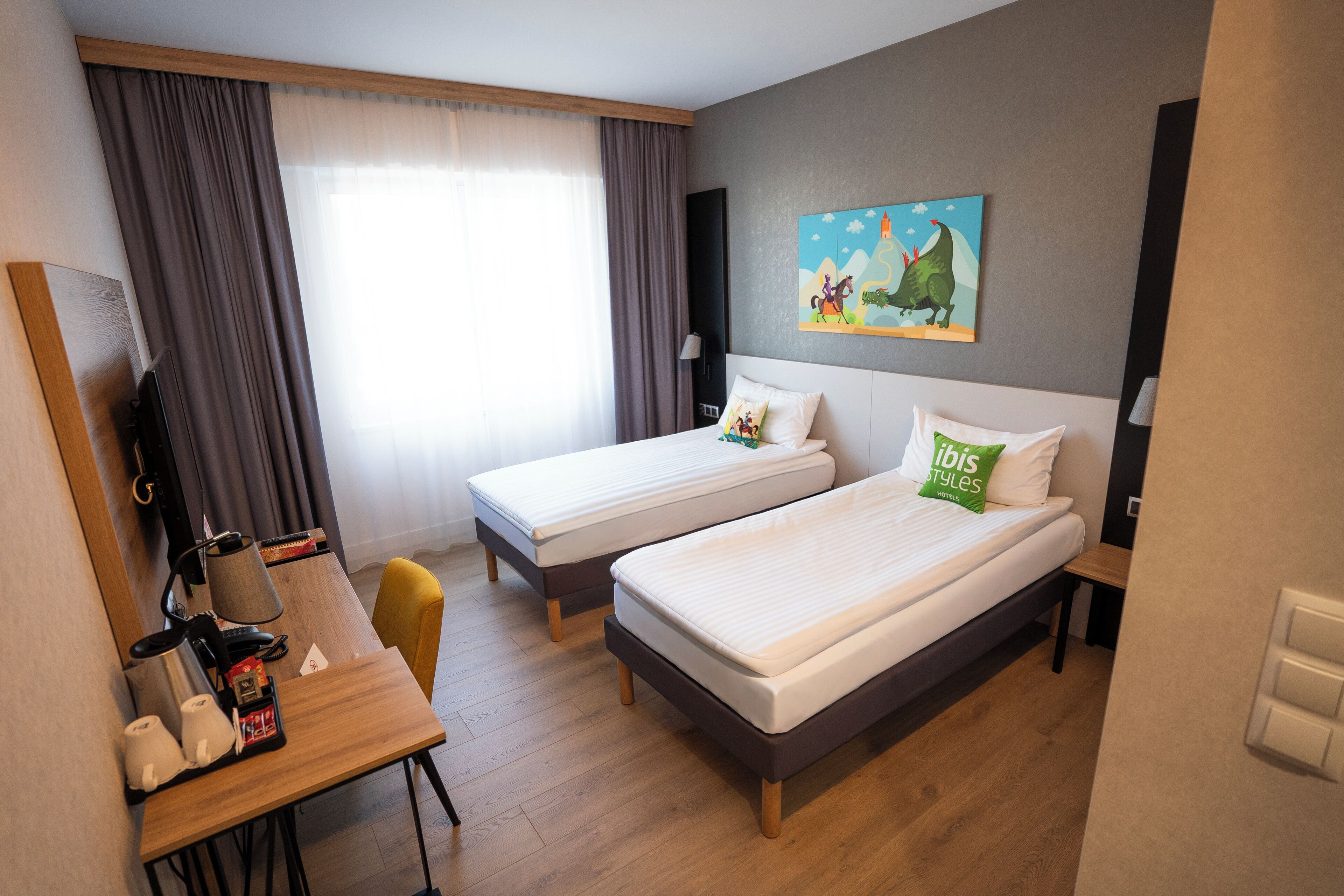 Ein Hotelzimmer mit zwei Einzelbetten, einem Schreibtisch mit Stuhl, einem Fernseher und einem Bild an der Wand.
