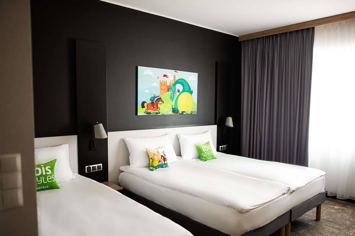 A hotel room with two single beds, a large wall-mounted painting, a window with curtains, and a bedside table with a lamp.