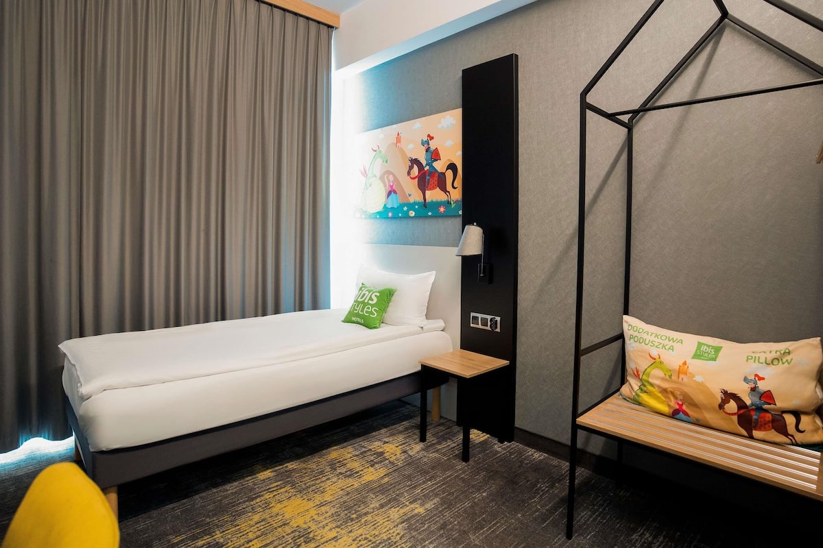 A hotel room with a bed, a nightstand, a wall-mounted lamp, a painting, and a hanging chair.