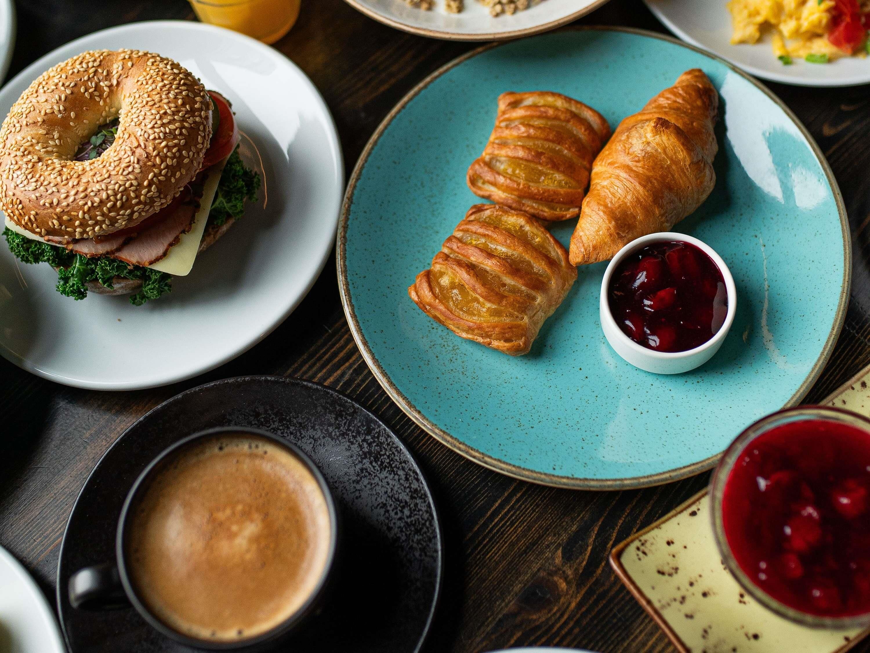 Uma mesa com um croissant, um sanduíche de bagel, uma xícara de café e um prato com uma pequena tigela de geleia.