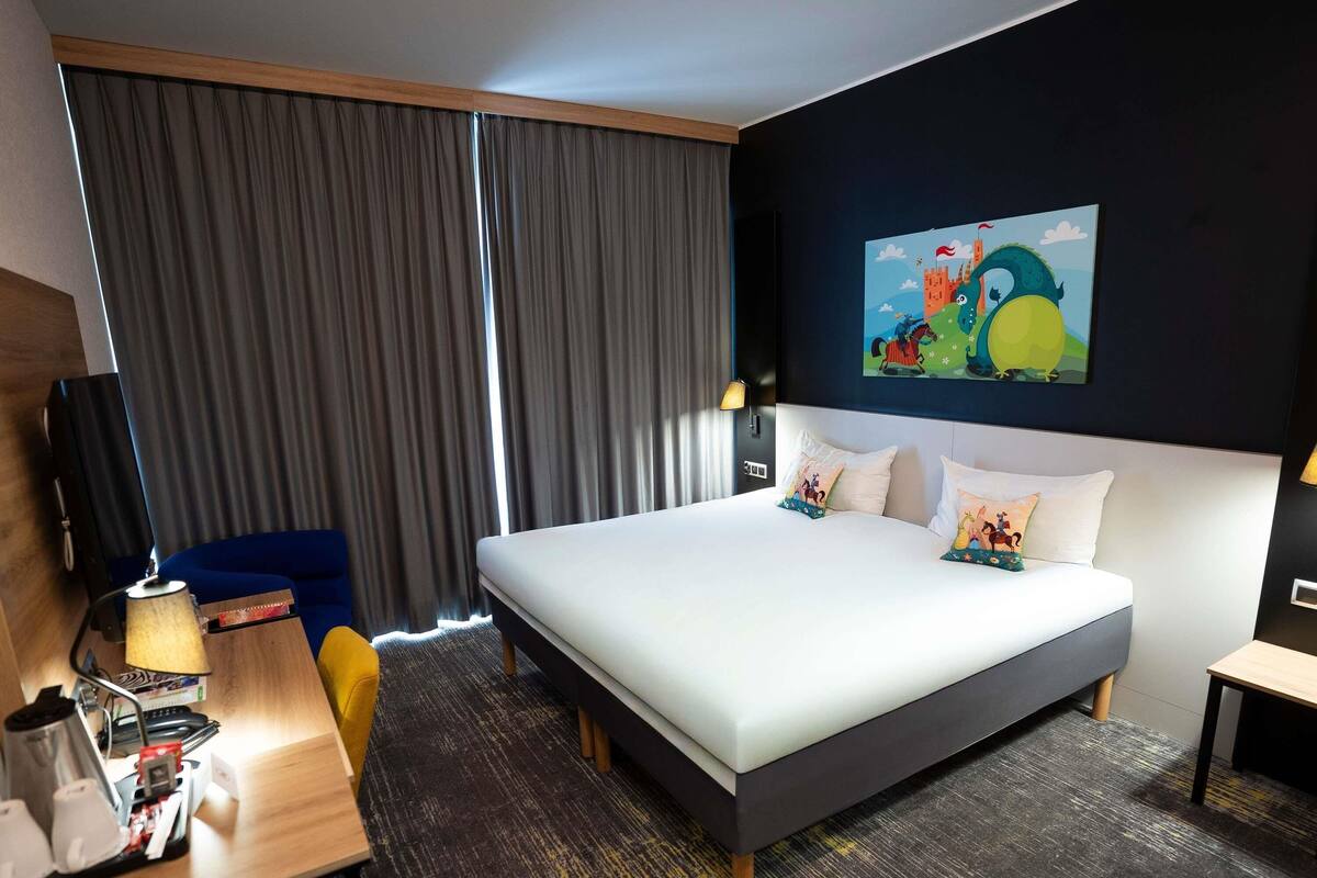 A hotel room with a large bed, a desk with a lamp, a chair, and a painting on the wall.