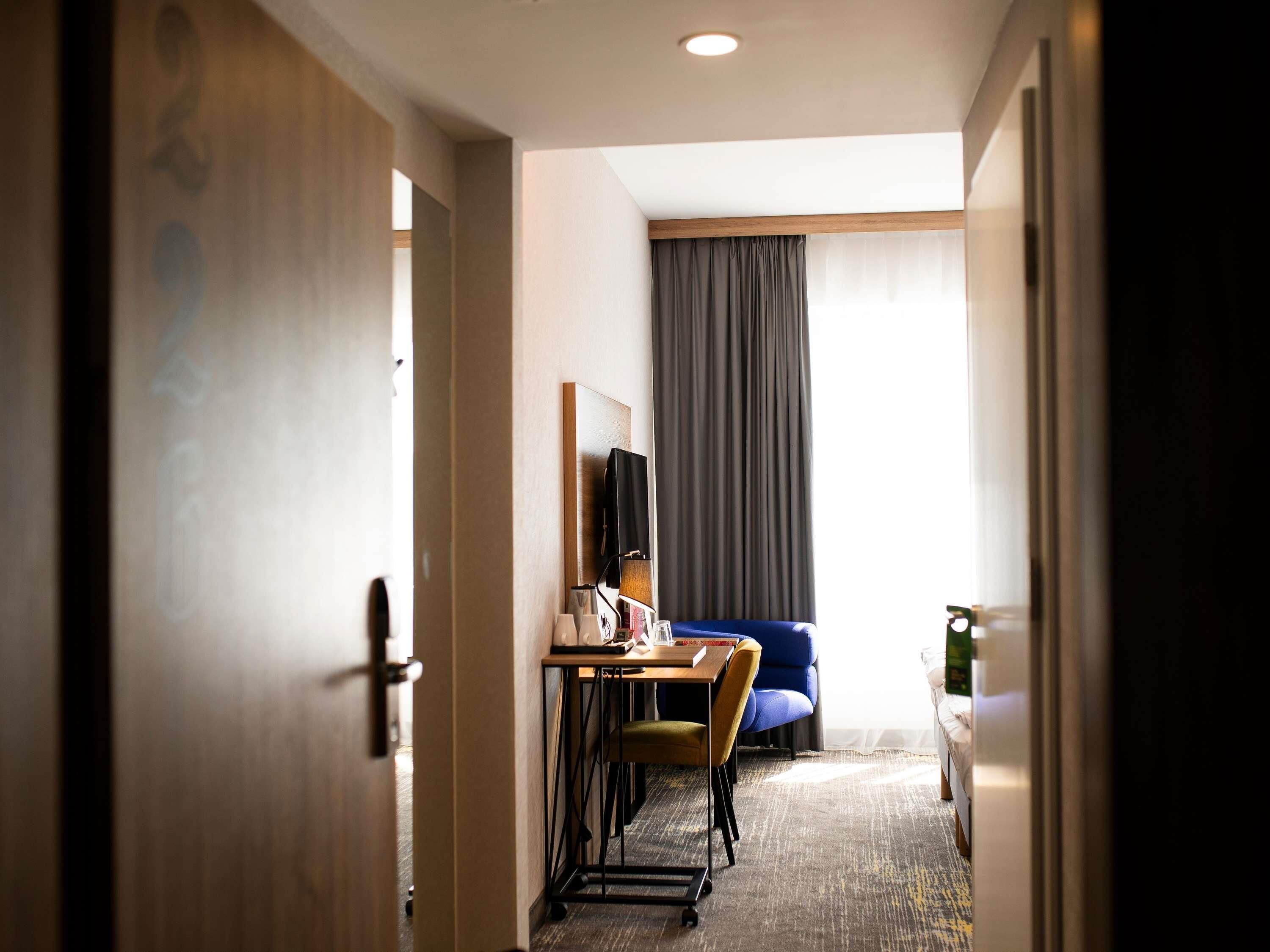 A hotel room with a desk, chairs, a bed, a mirror, and a window with curtains.