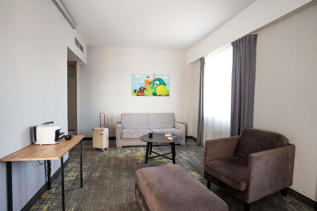 A hotel room with a sofa, armchair, ottoman, coffee table, and a small table with a coffee maker.