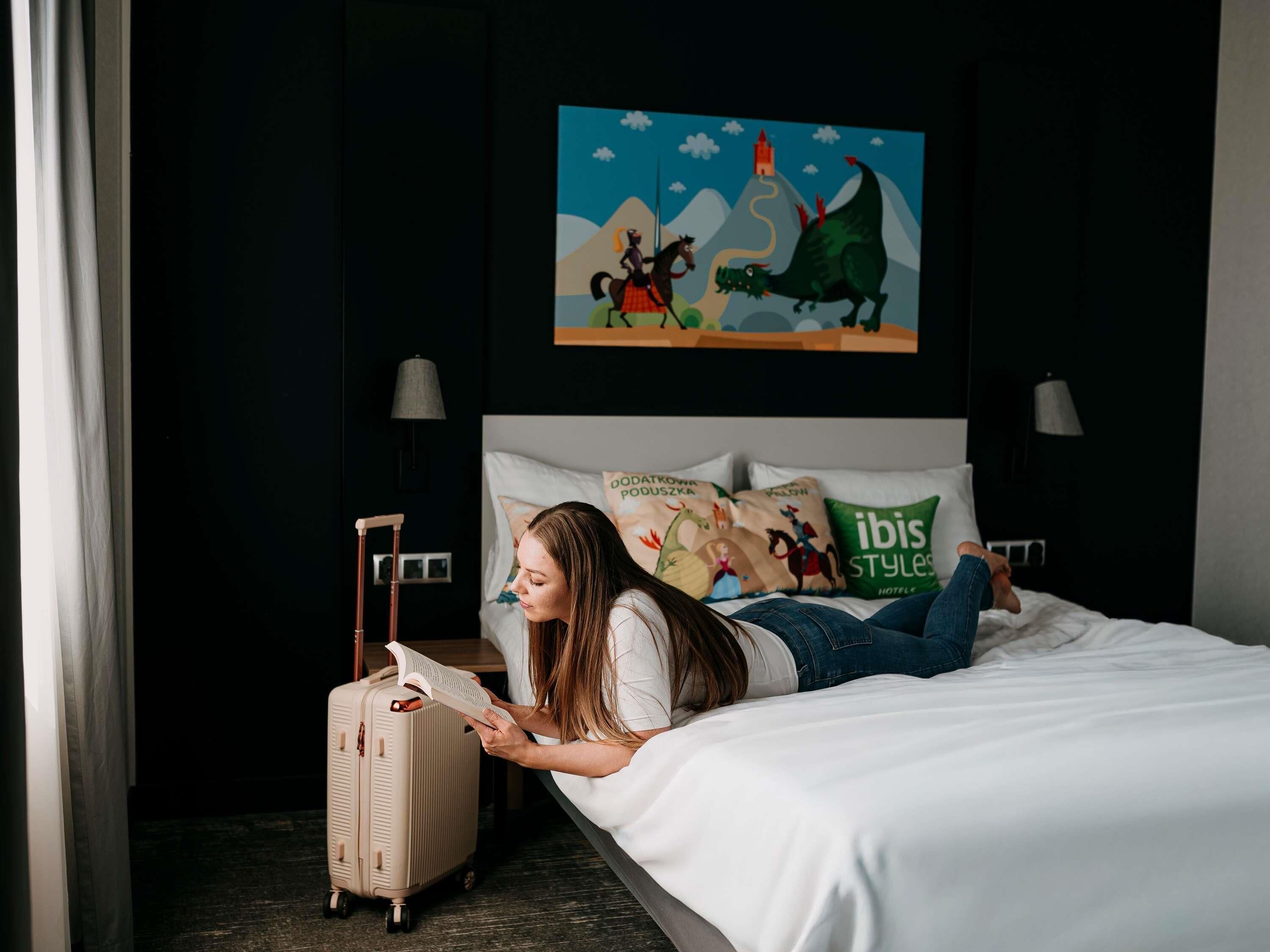 A person lying on a bed reading a book, with a suitcase beside them, in a room with a painting on the wall and pillows with text.
