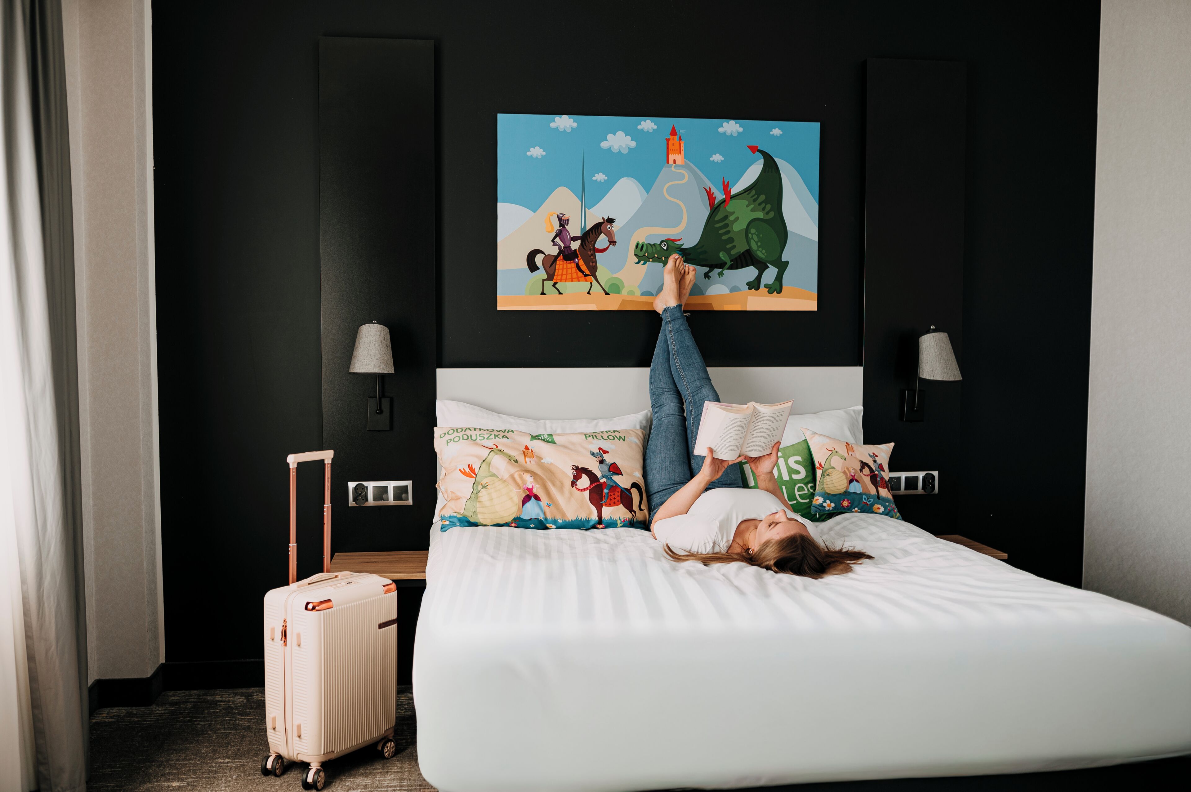 A bed with a colorful pillow, a suitcase, and a painting of a horse and dinosaur on the wall.