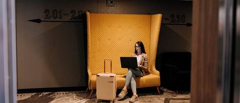 A person sitting on a yellow cushioned chair with a laptop, a suitcase, and a wall with room numbers 201-230.
